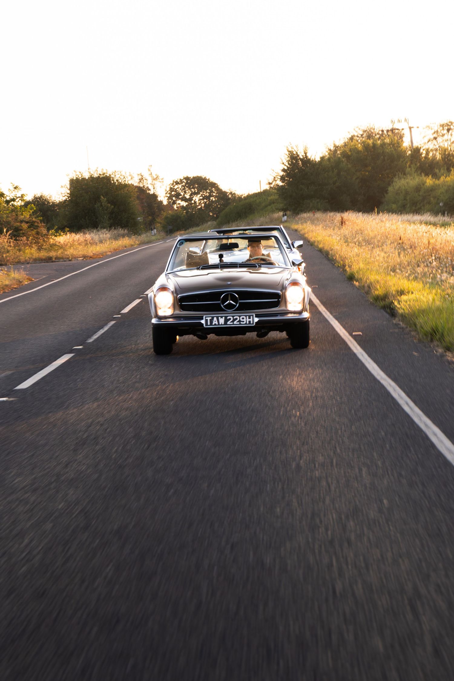 Classic Mercedes-Benz convertible driving on a rural road at golden hour sunset.