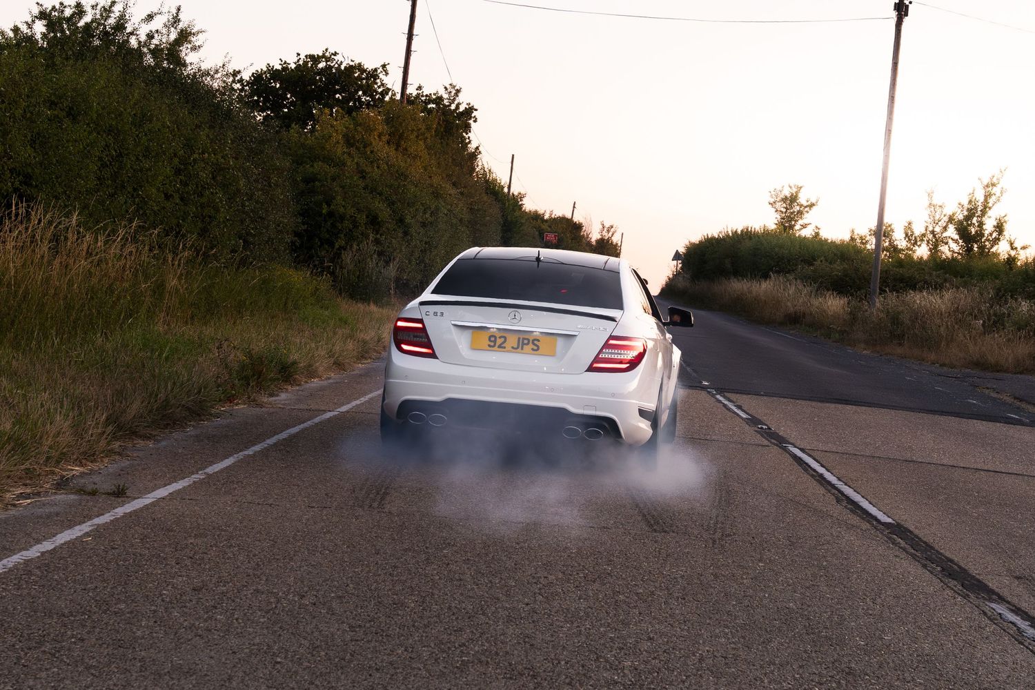 White sedan performing a burnout on a rural road, with tire smoke billowing from the rear wheels at dusk.