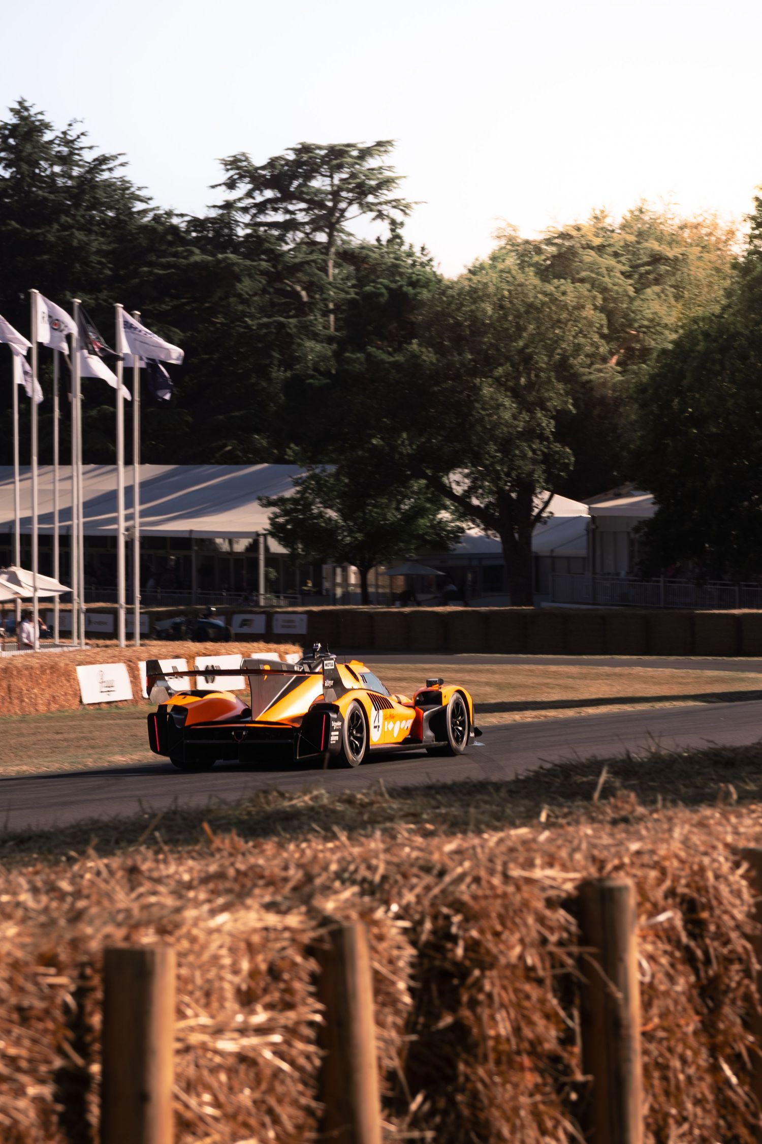 Orange McLaren race car on track at Goodwood Festival of Speed, with flags and trees in background.