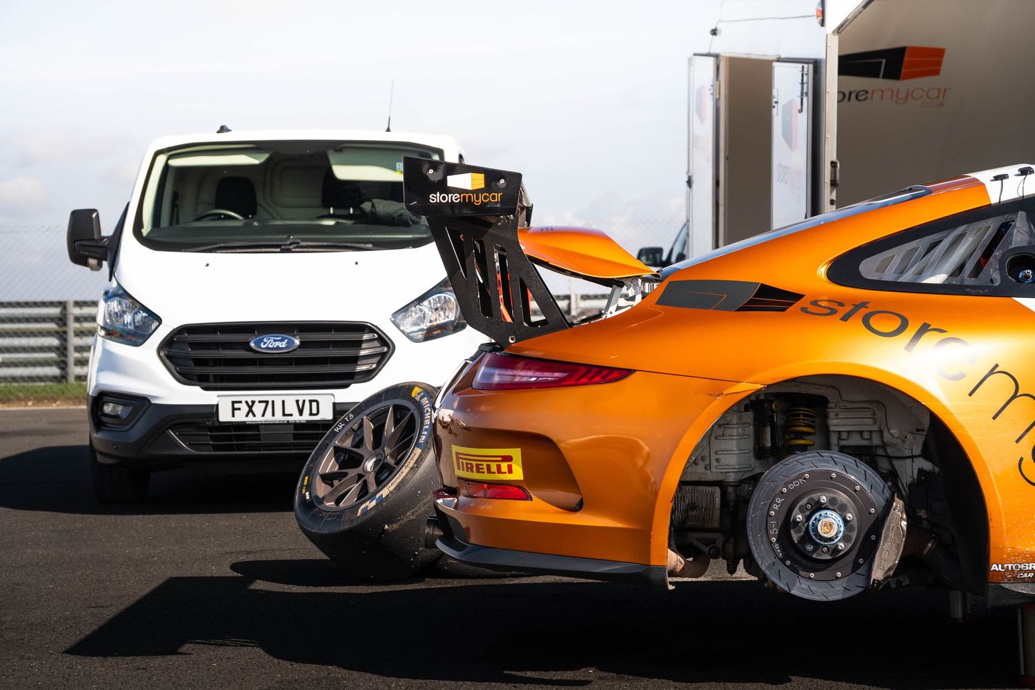 Orange Porsche 911 GT3 race car rear wing detail alongside a Ford Transit van at a circuit.