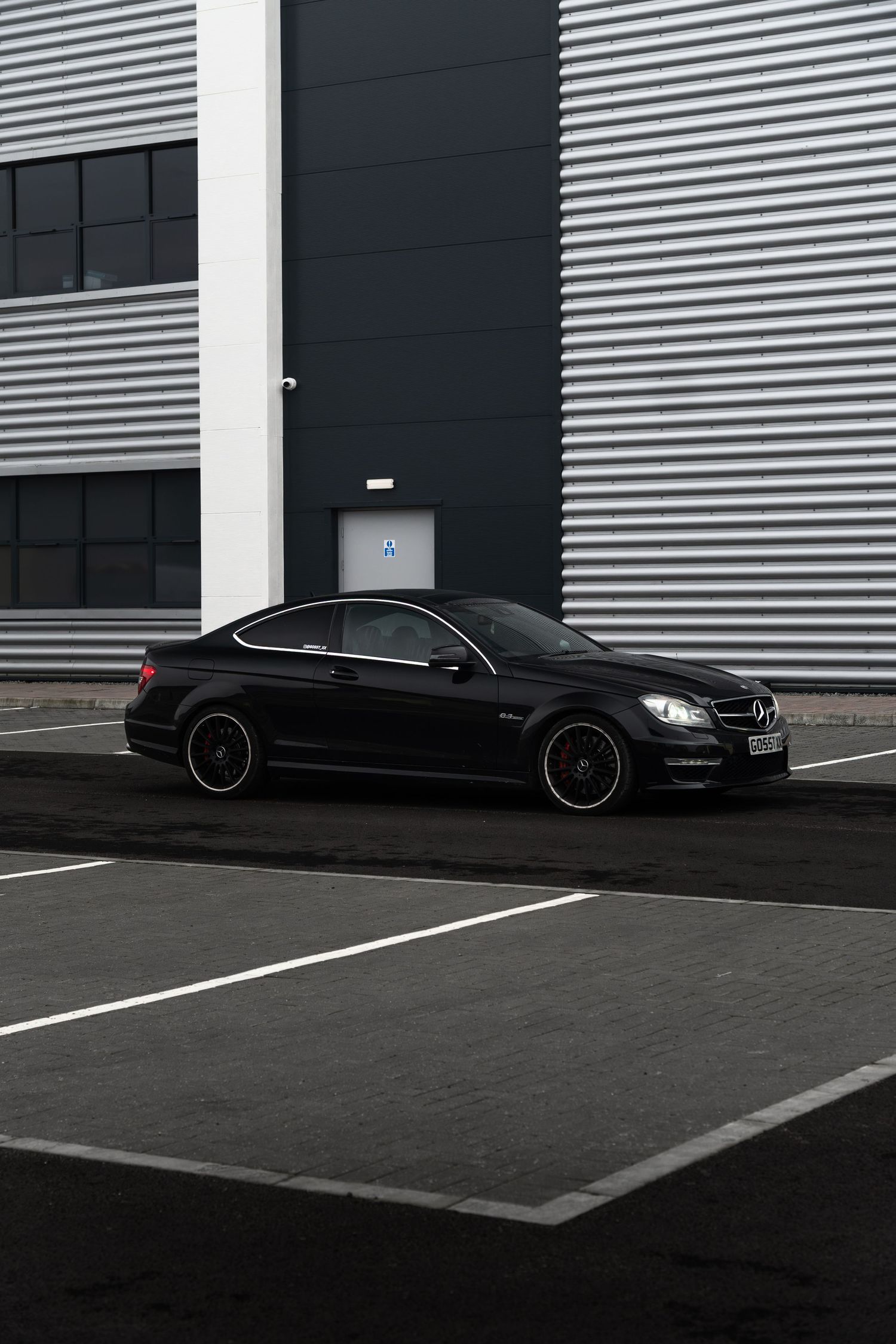 Black Mercedes-Benz C63 AMG coupe parked outside modern industrial building with security camera.