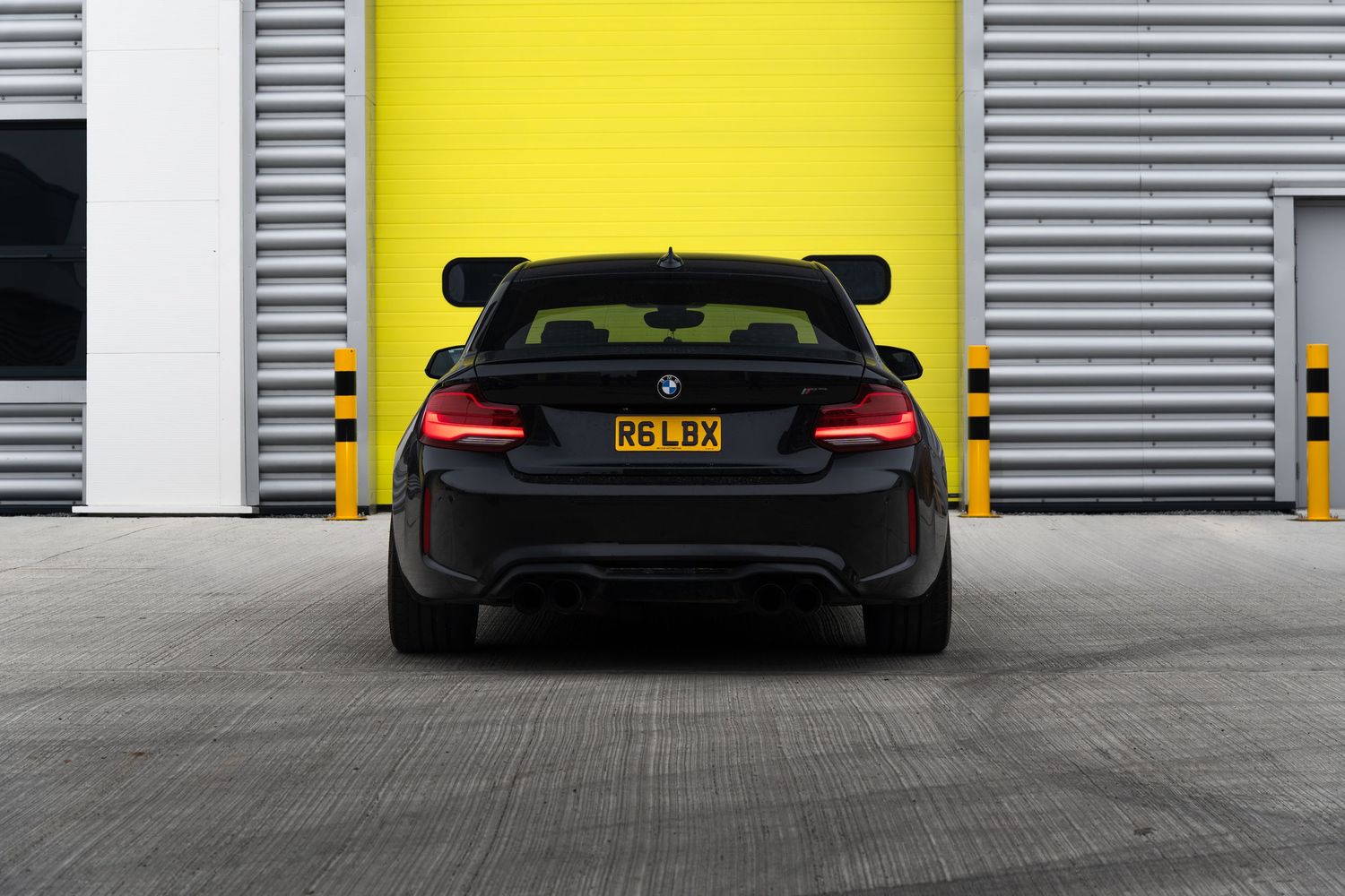 Rear view of a black BMW M2 coupe with plate R6LBX parked before yellow garage doors.