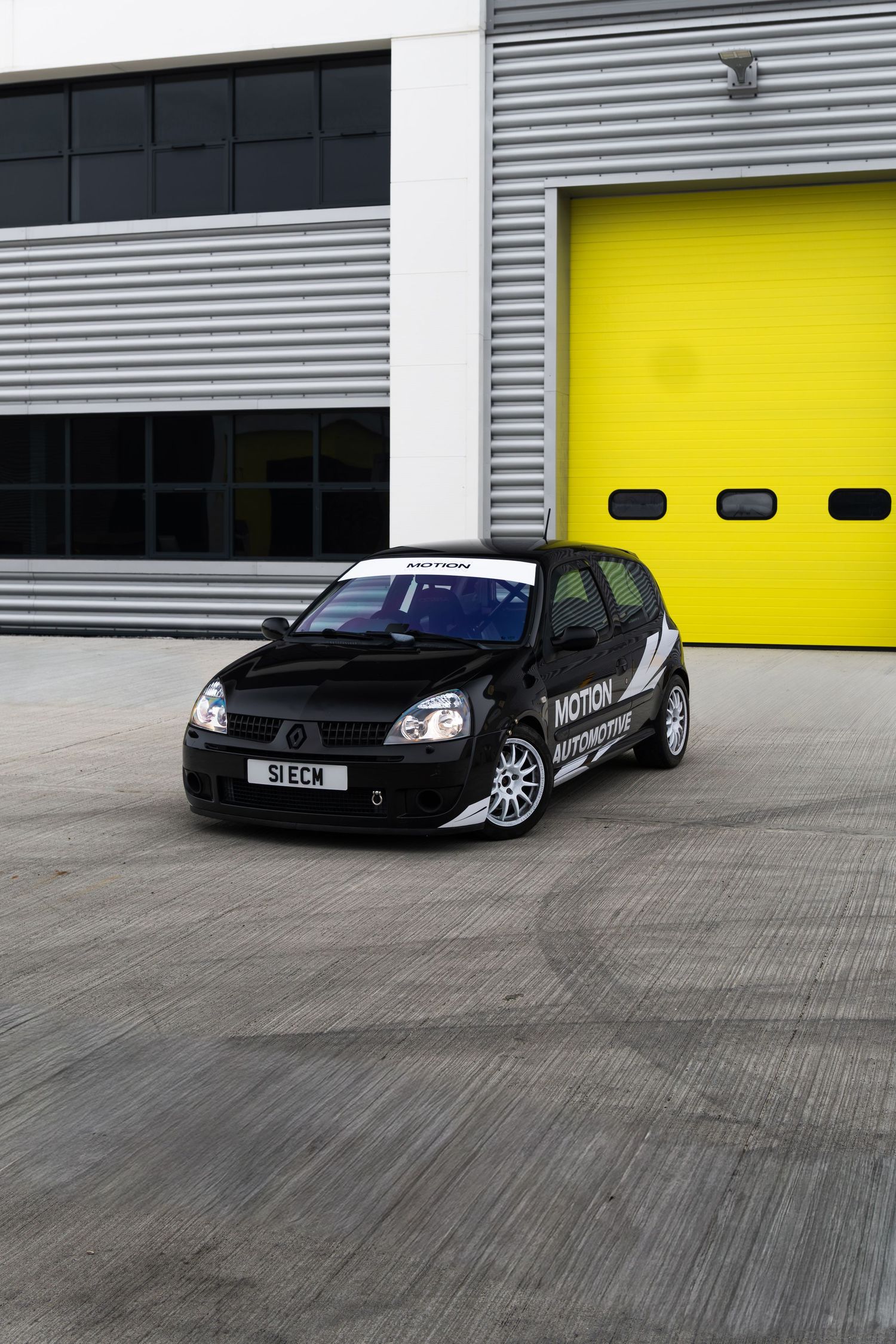 Black Renault Clio with Motion motorsport graphics parked outside industrial unit with yellow doors.