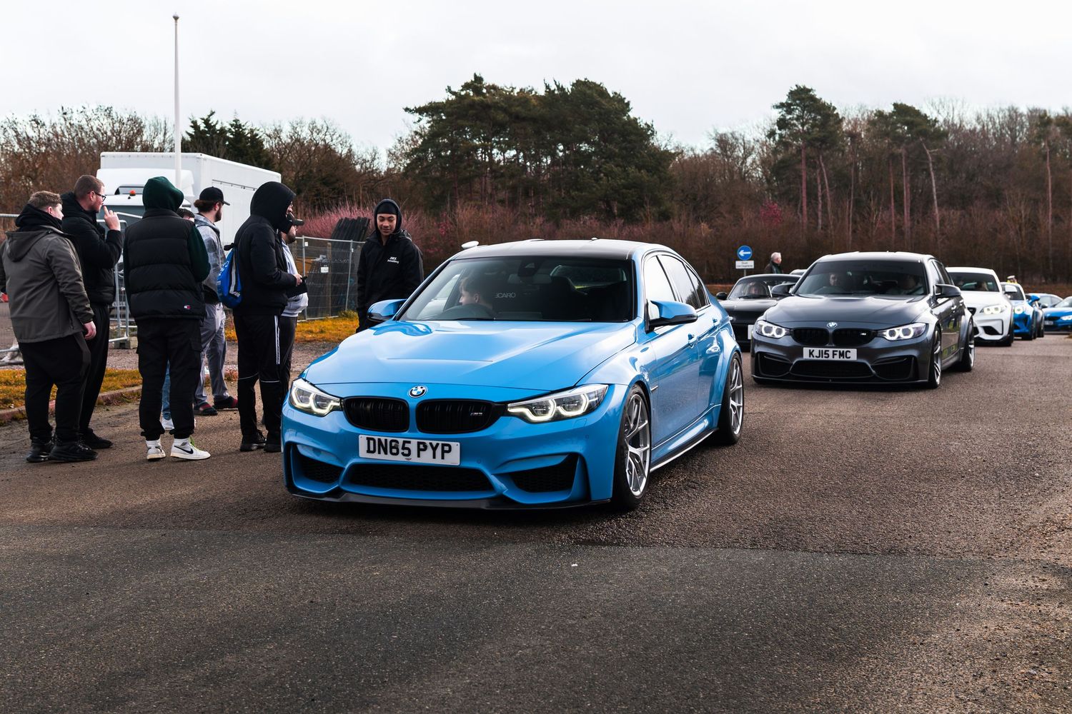 A blue BMW M3 leads a row of performance cars at an outdoor car meet event.