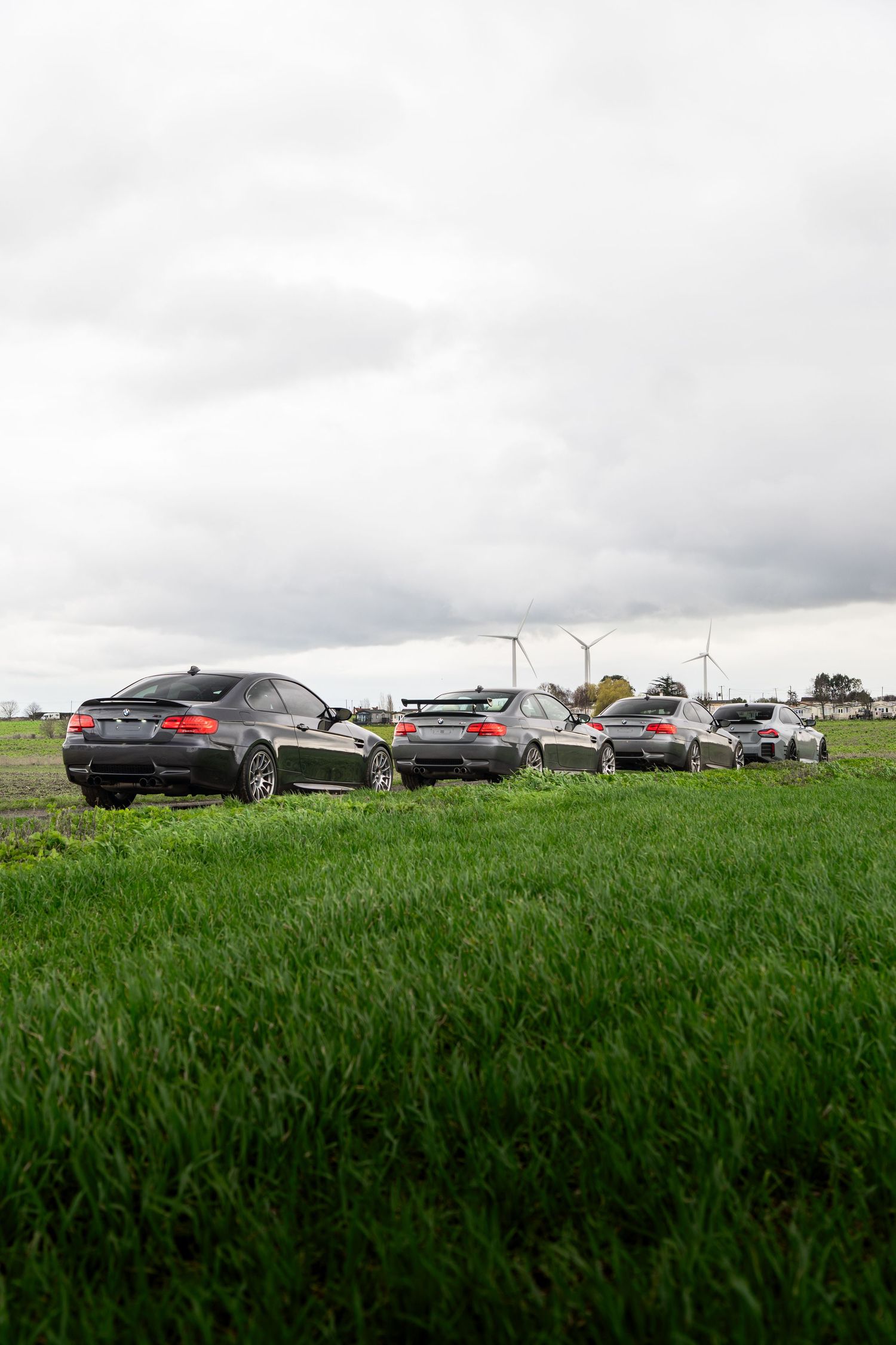 Multiple dark BMW M3 coupes lined up in green field with wind turbines visible.