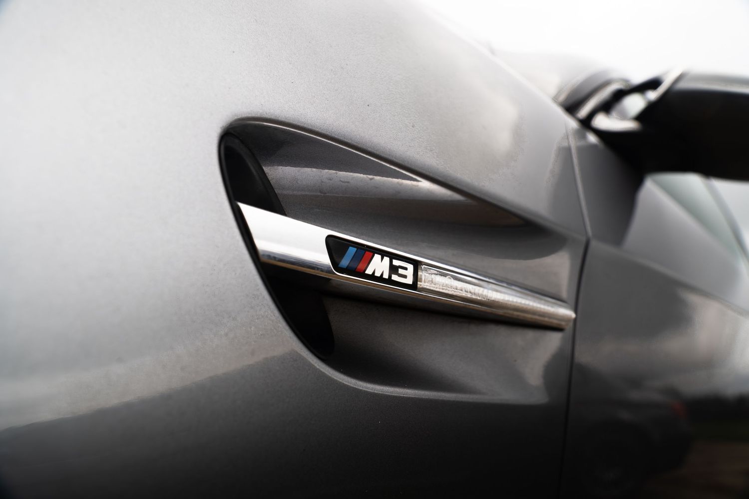 Close-up of BMW M3 fender badge with iconic M tricolor logo on dark gray body panel.