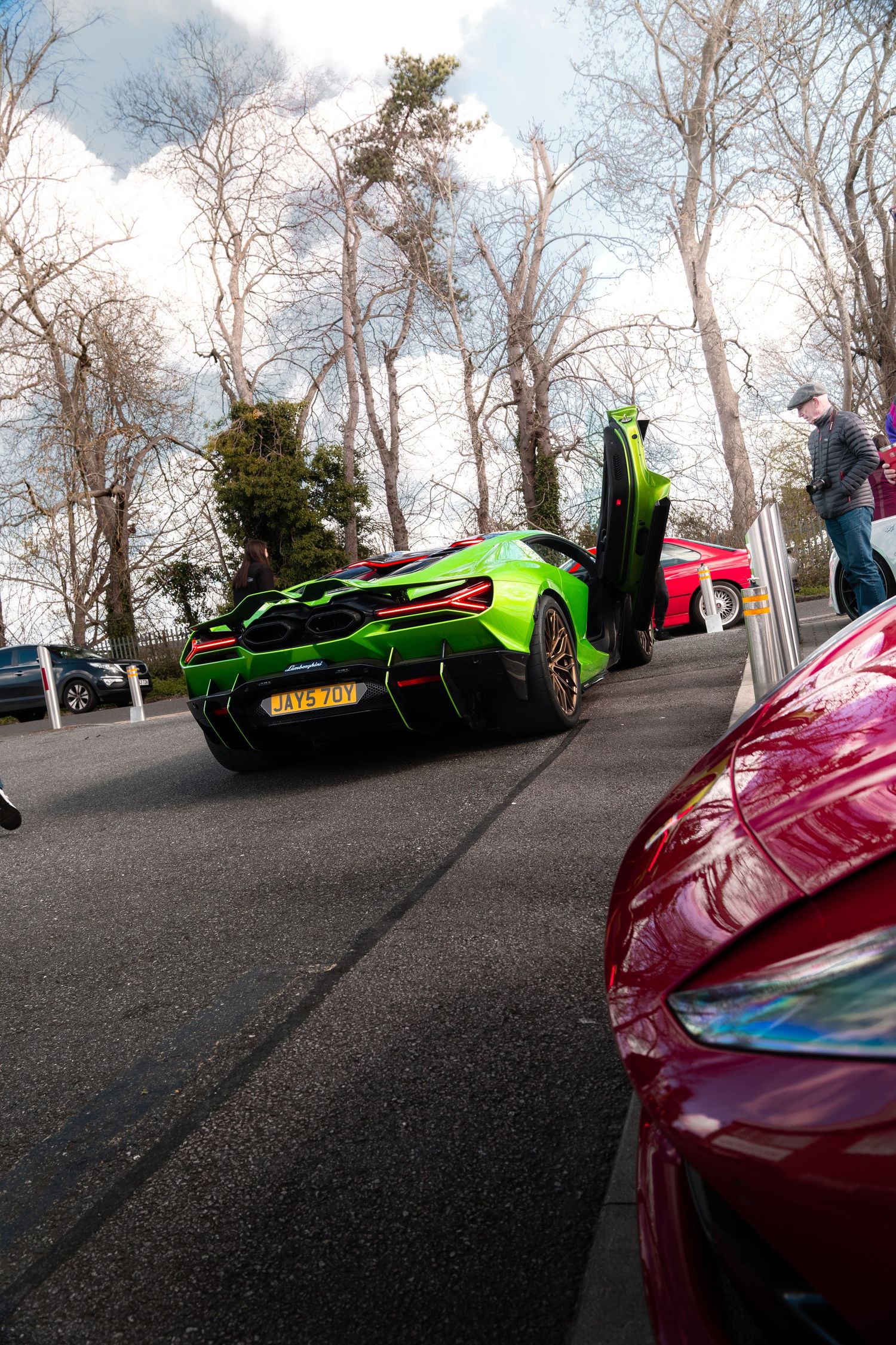 Lime green McLaren supercar with dihedral doors open in a parking lot surrounded by exotic cars and bare trees.