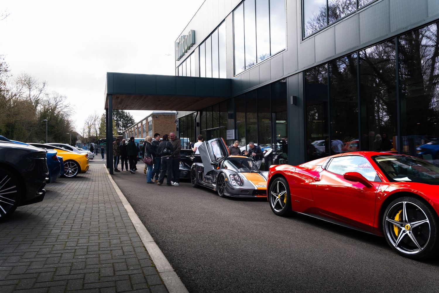 Row of exotic supercars including red Ferrari 458 and Pagani outside modern dealership building.