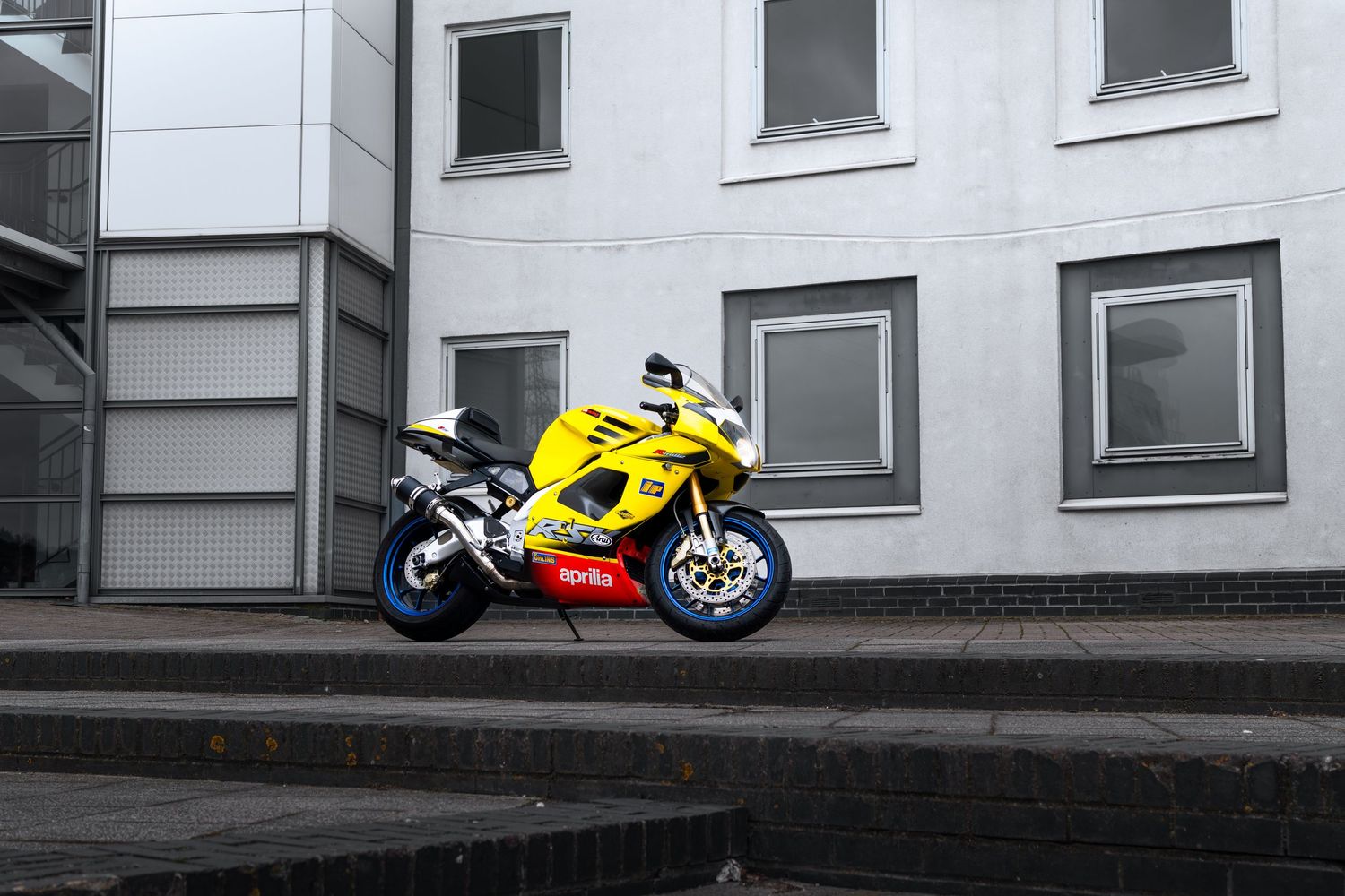 Yellow Aprilia RSV1000 sportbike parked on stepped pavement in front of a modern urban building.