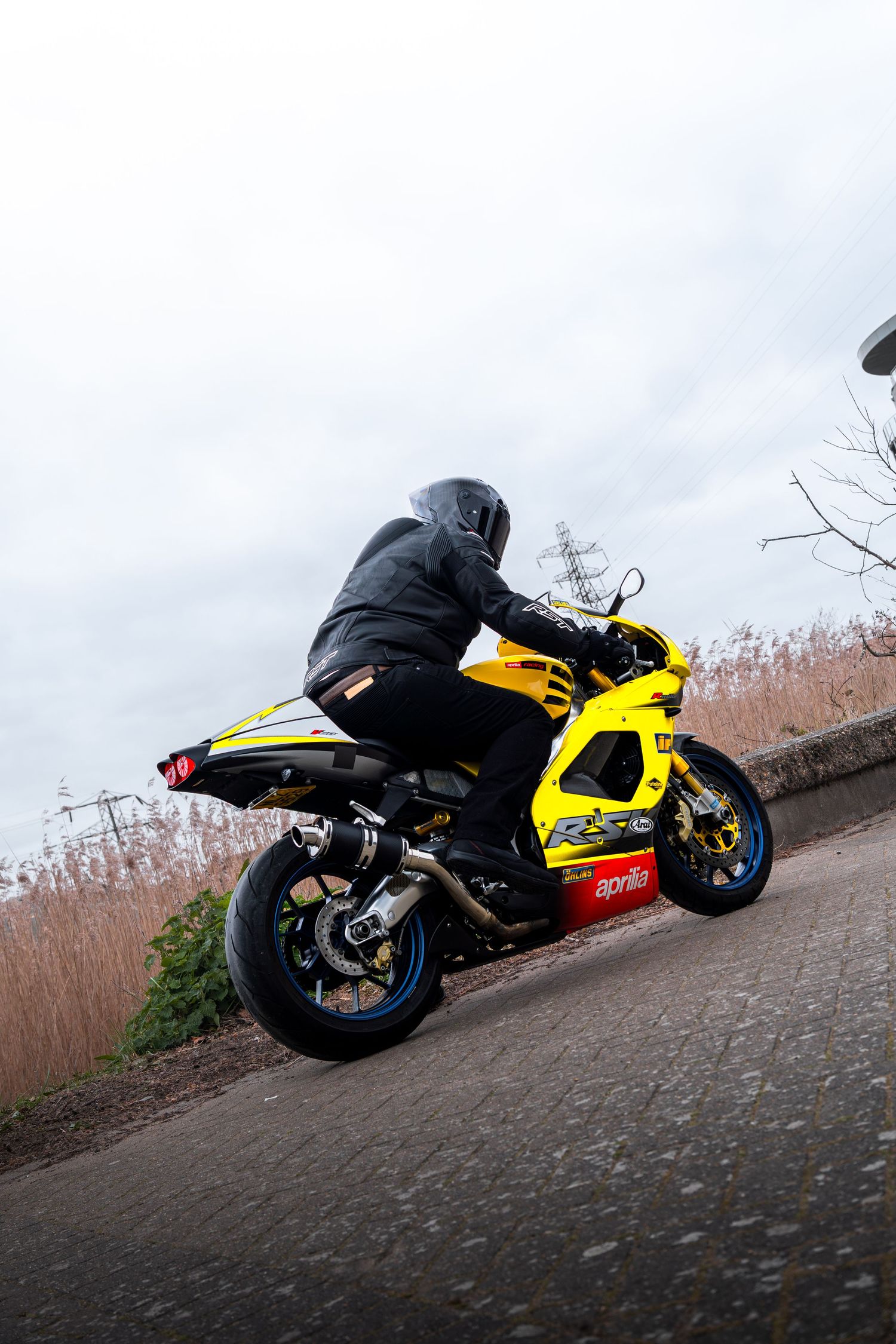 Rear three-quarter view of a yellow Aprilia sportbike with black-clad rider on a road.