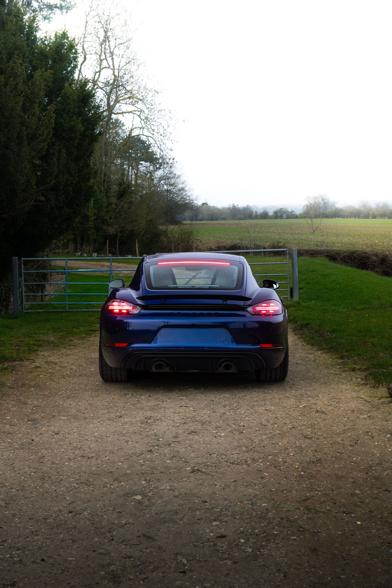 Blue Porsche 718 Cayman rear view on a gravel driveway with a metal gate and green fields.