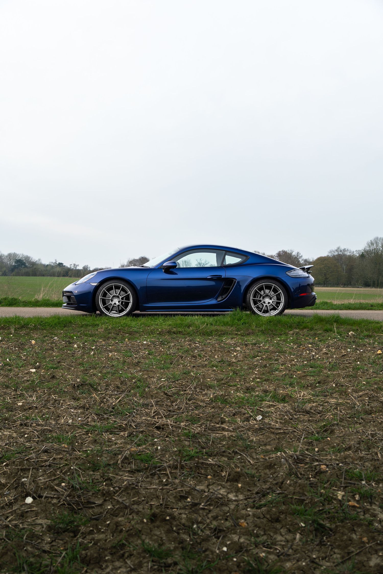 Deep blue Porsche 718 Cayman GTS parked on a rural road beside a green field under an overcast sky.