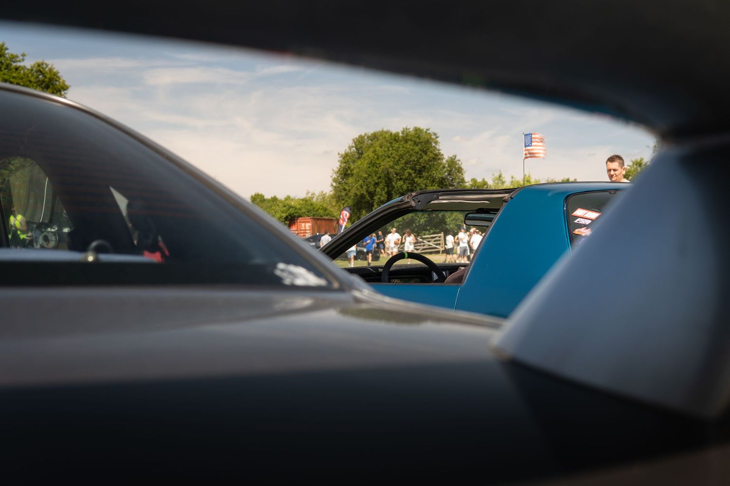Close-up view between luxury sports cars at an outdoor car meet on a sunny day.