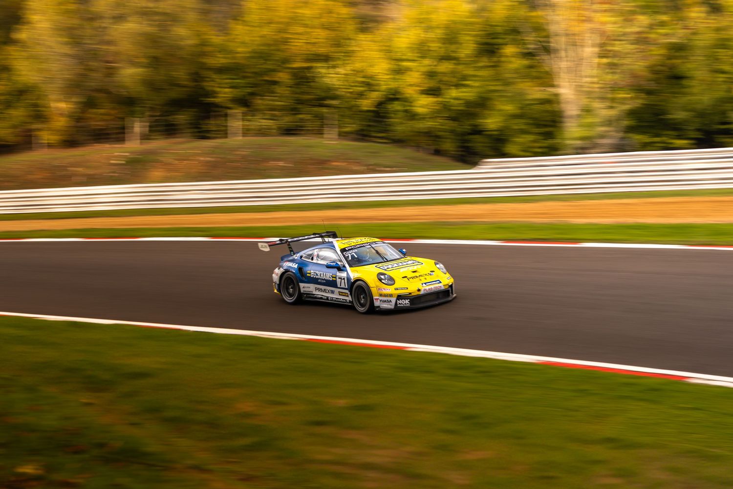 Yellow and blue Porsche 911 GT3 race car navigating a circuit with colorful autumn foliage backdrop.