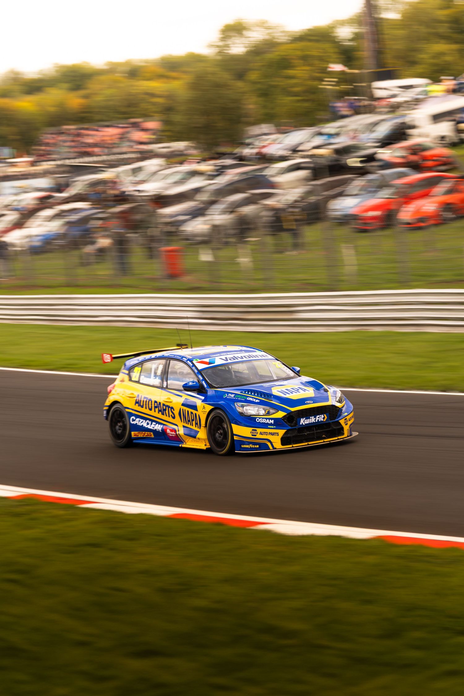 A yellow and blue Ford Focus BTCC race car speeds through a corner at a motorsport circuit.