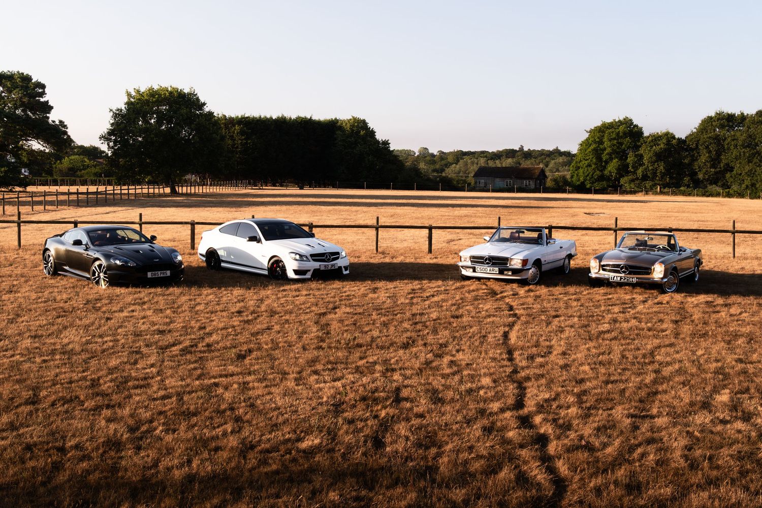 Four classic and modern Mercedes-Benz cars parked in a golden field with trees and fencing in the background.