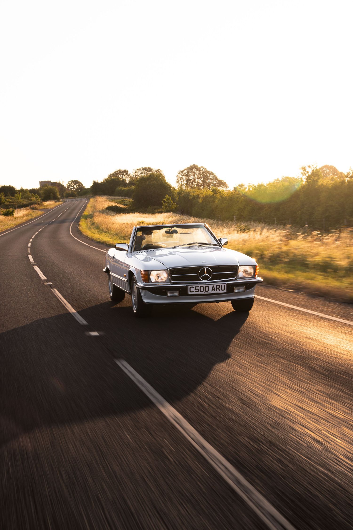 Classic Mercedes-Benz convertible driving on a rural road at golden hour sunset in the UK.