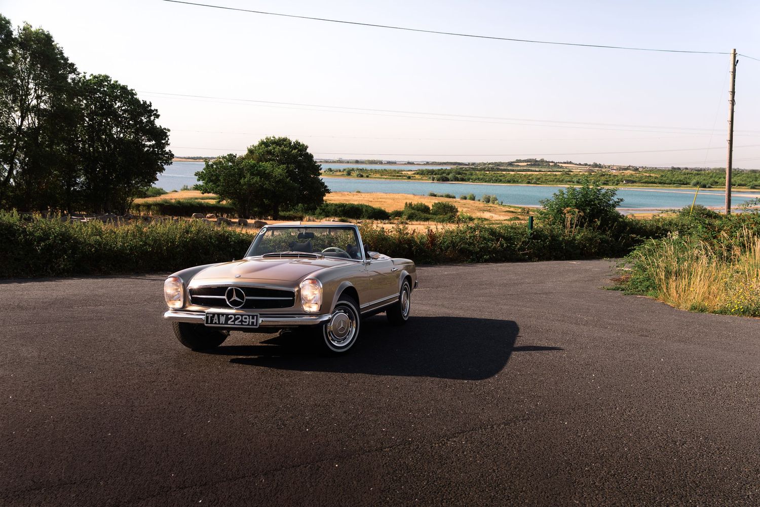 Classic silver Mercedes-Benz 280SL convertible parked on a country road with scenic water and green fields in background.