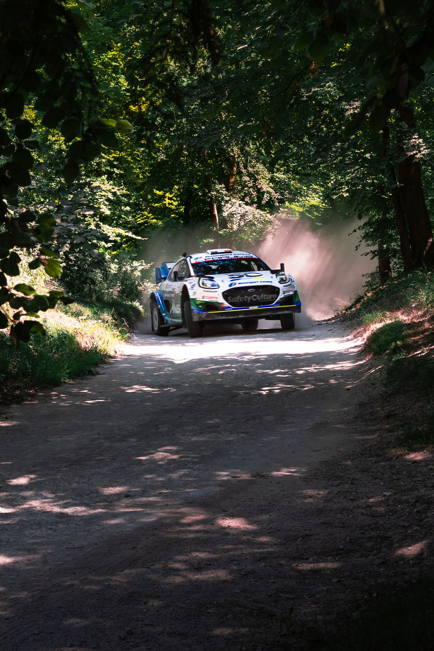 Rally car racing through a dusty forest road surrounded by lush green trees during a daytime rally event.