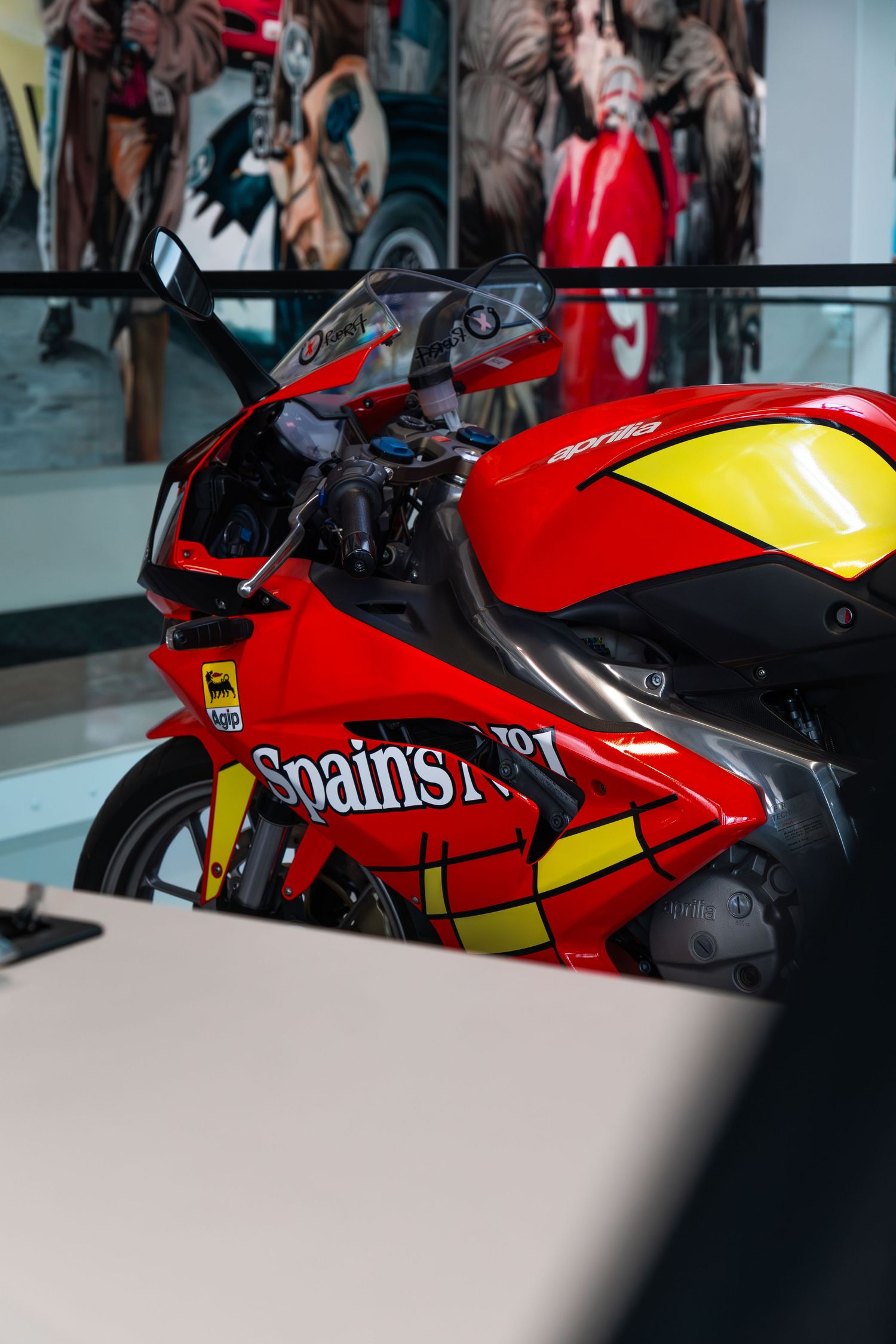Red and yellow Aprilia Spain's No.1 racing motorcycle displayed indoors with colorful mural backdrop.