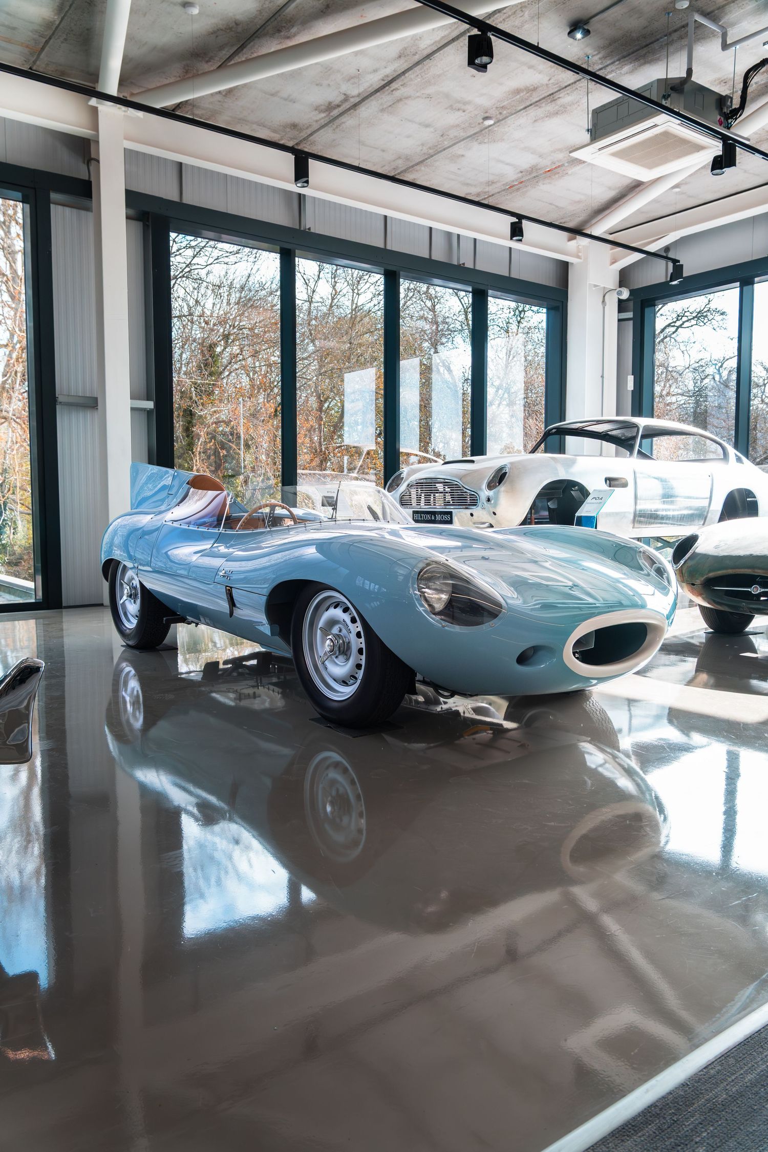 Light blue vintage Jaguar D-Type racing car displayed on a polished showroom floor with large windows.