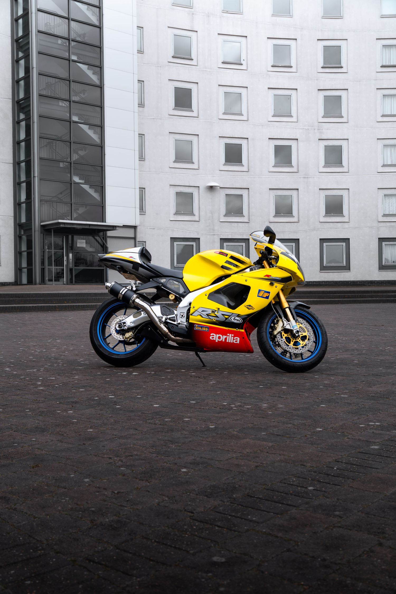 Yellow and red Aprilia RSV sportbike in profile view parked on cobblestones before a white building.
