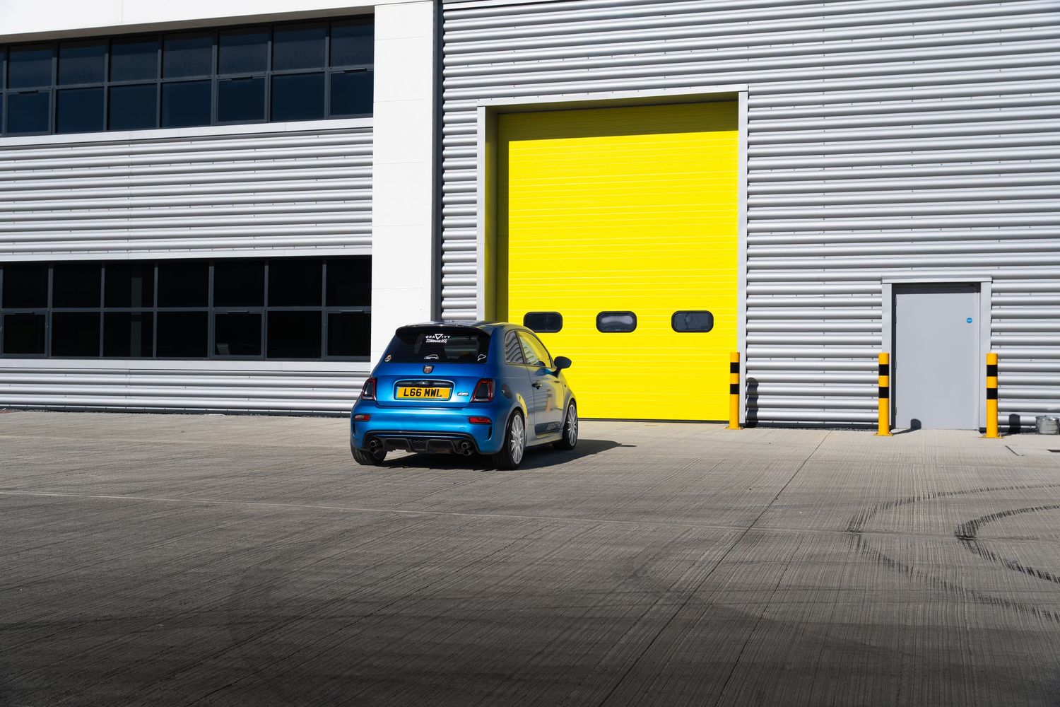 Blue Abarth 595 hatchback parked on concrete apron beside large yellow roller shutter doors.