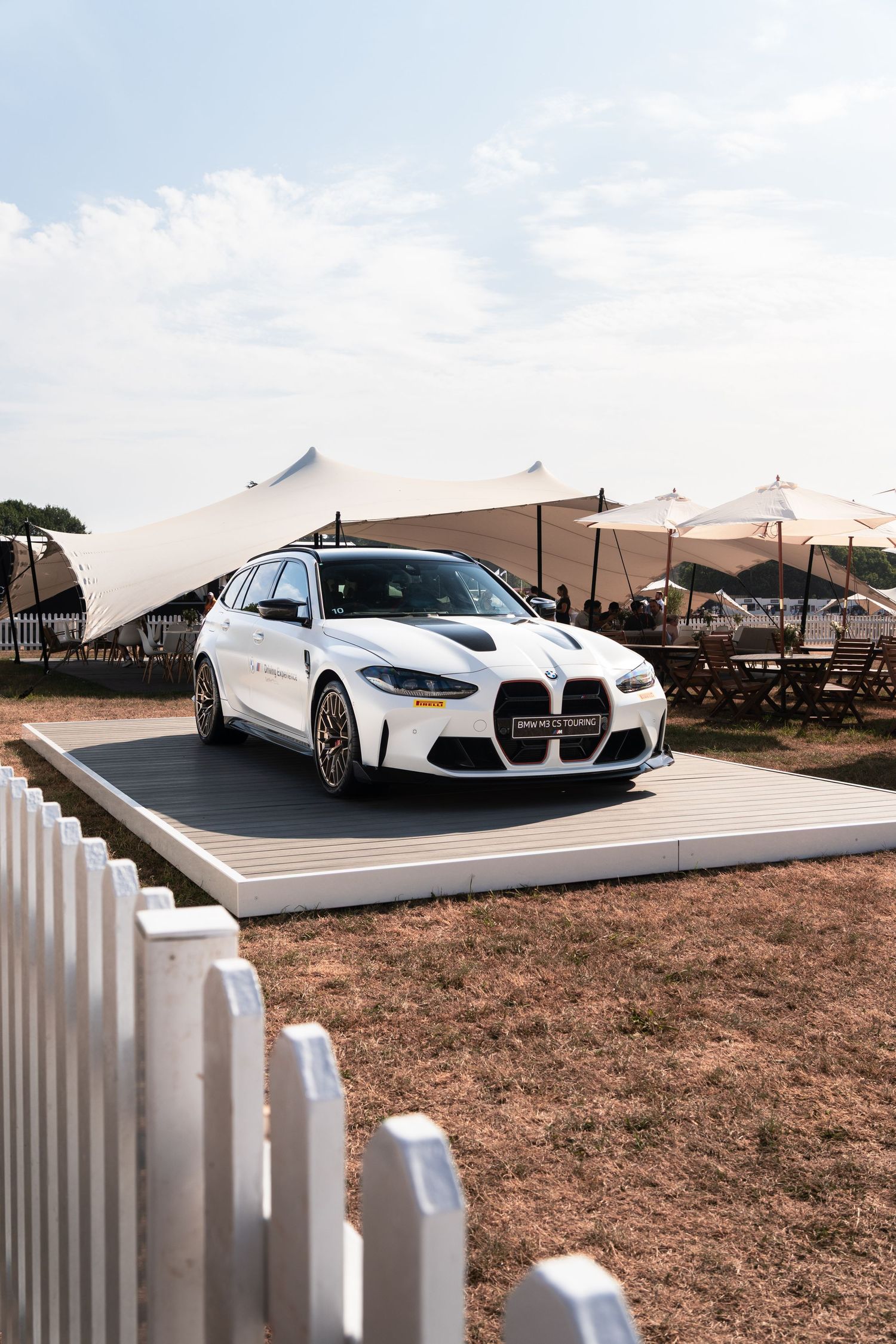 White BMW M3 Touring wagon on display platform at outdoor automotive event with tent canopy backdrop.