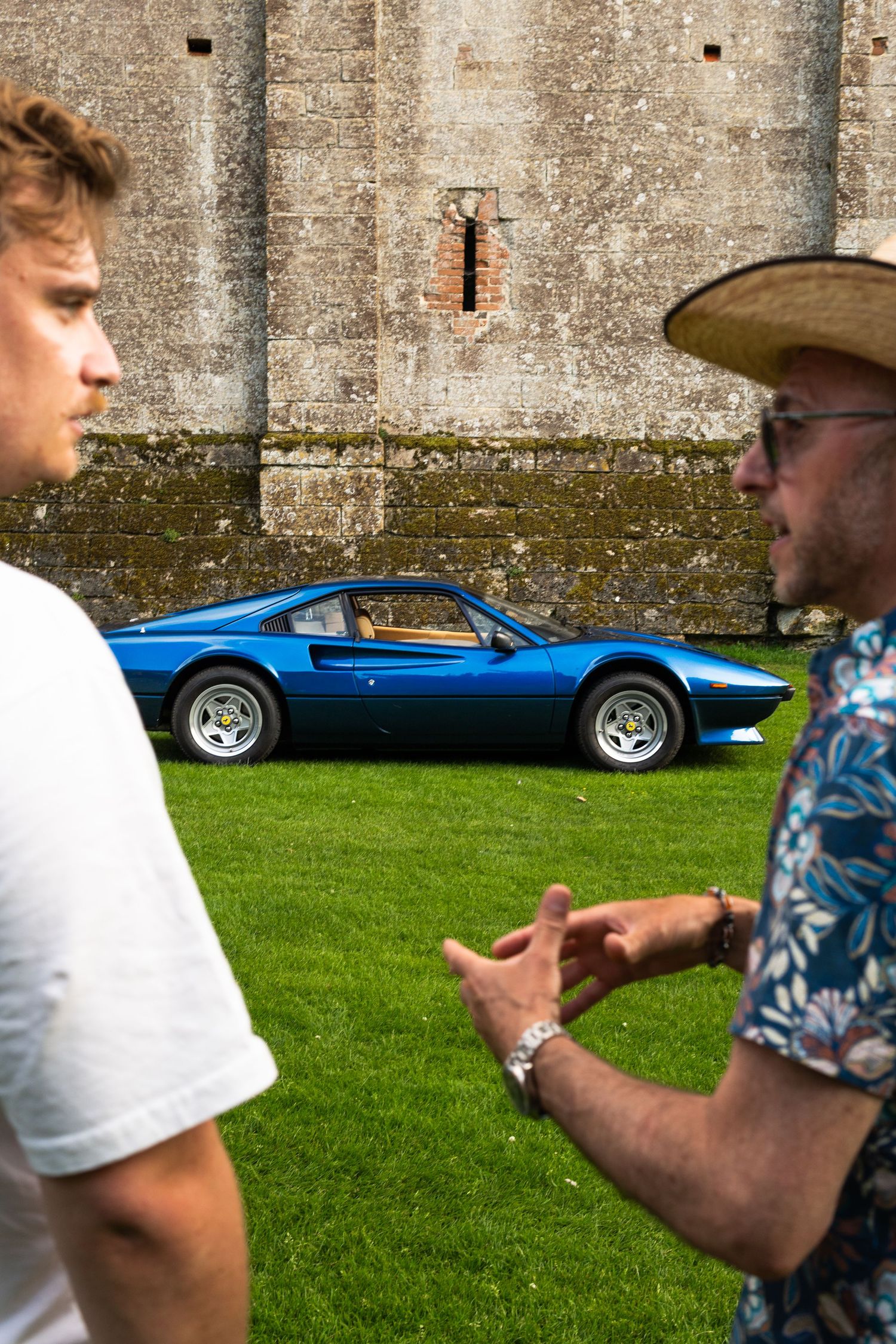 Two men discussing a classic blue Ferrari 308 GTB on green grass beside a stone wall.