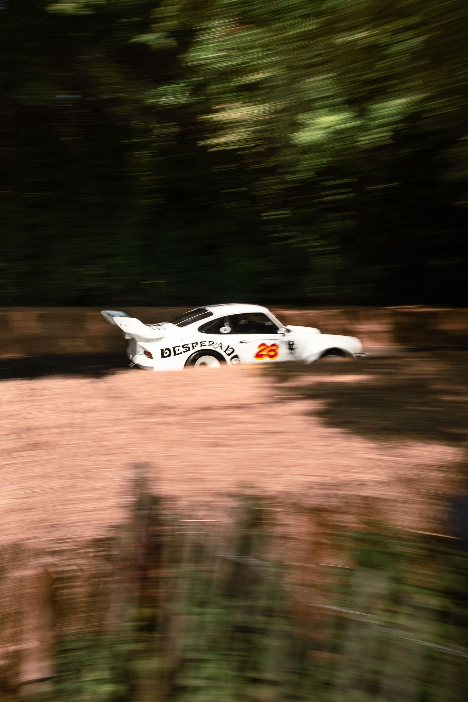 White race car number 28 with Desperado livery speeding through a dirt road surrounded by trees.