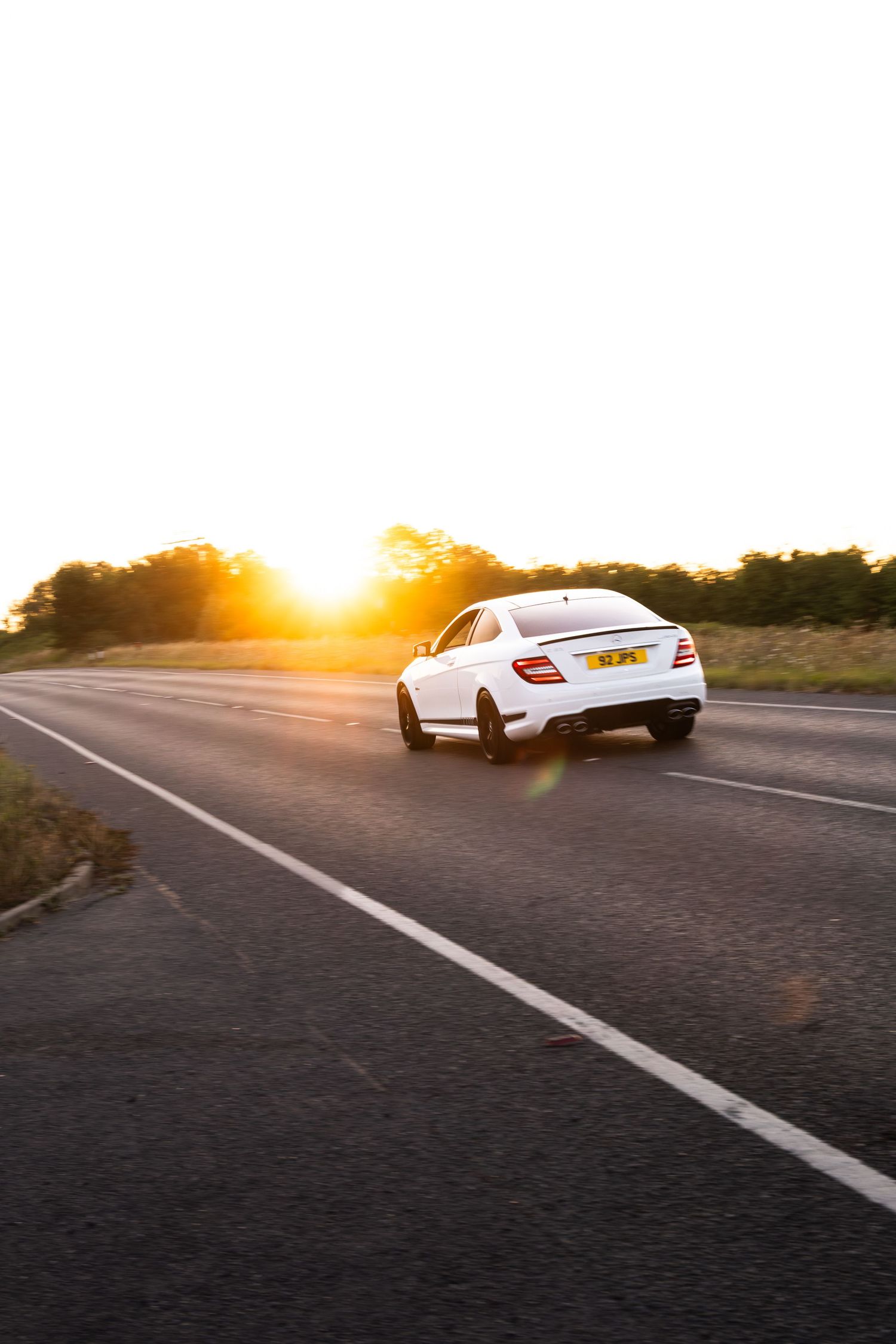 White Mercedes-Benz coupe driving on open road at sunset with golden light and tree line in background.
