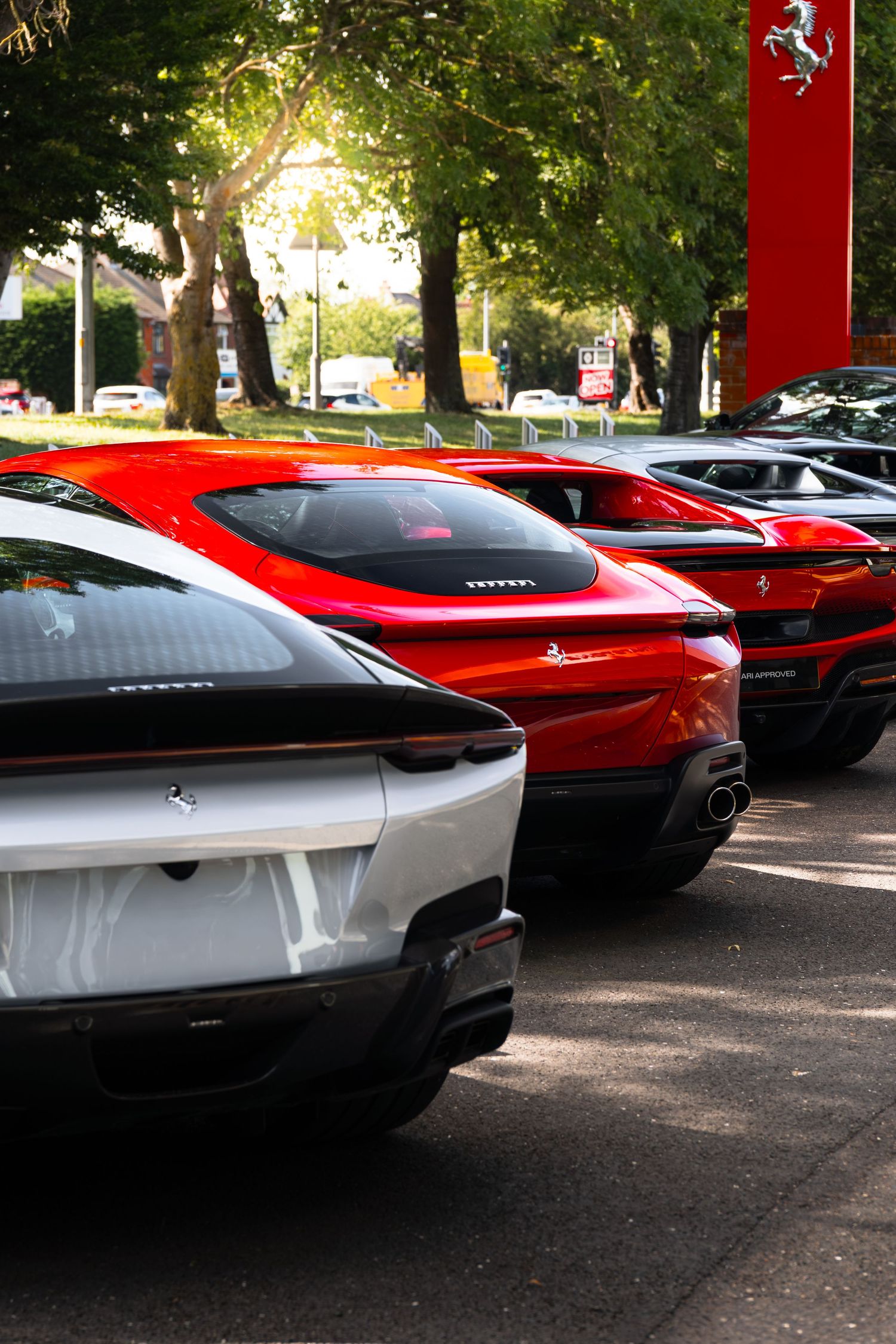 Row of Ferrari sports cars parked at a Ferrari dealership, including white and red models, with trees in background.