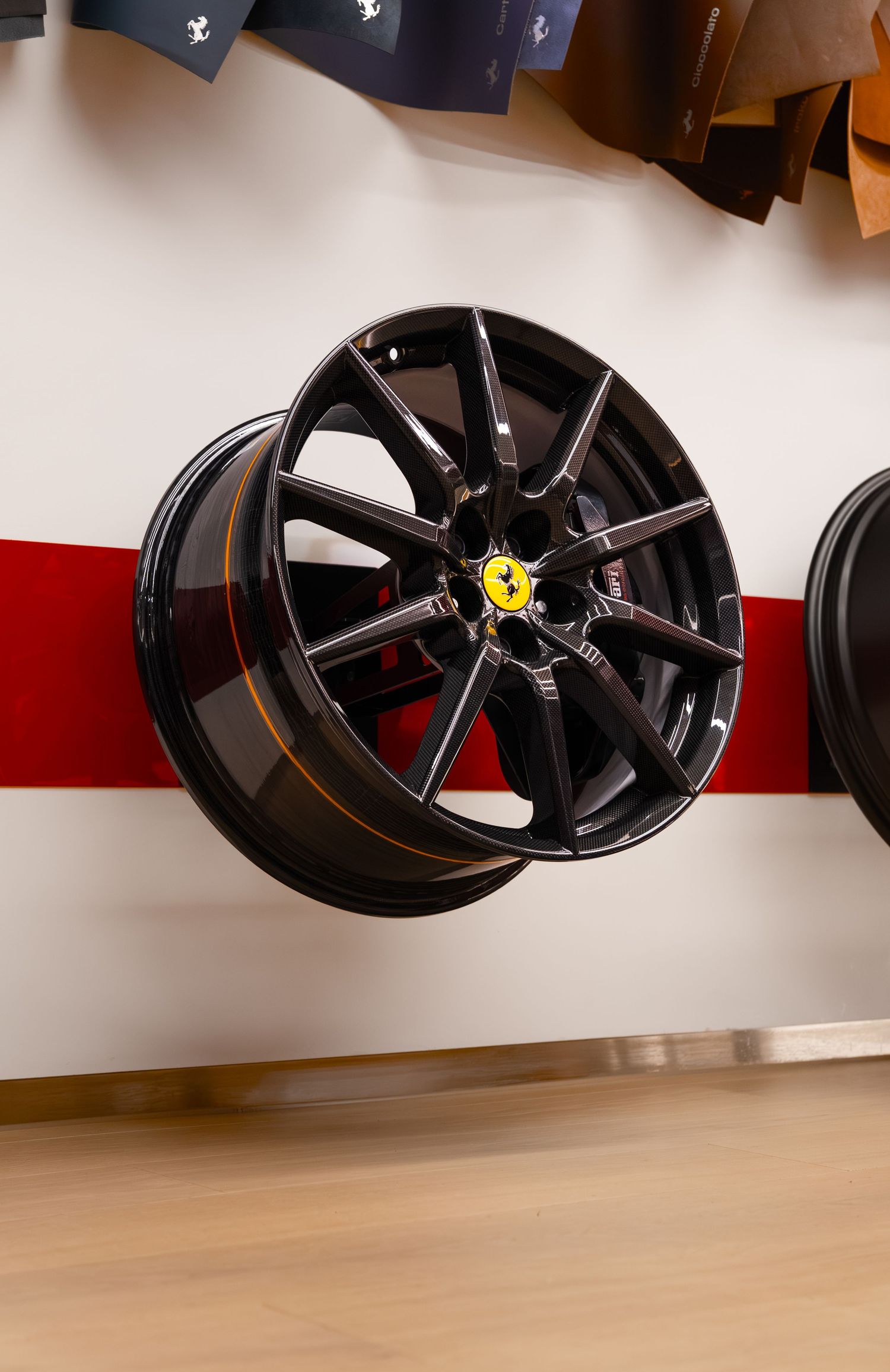 Glossy black Ferrari alloy wheel with yellow prancing horse logo displayed on a white and red showroom wall.