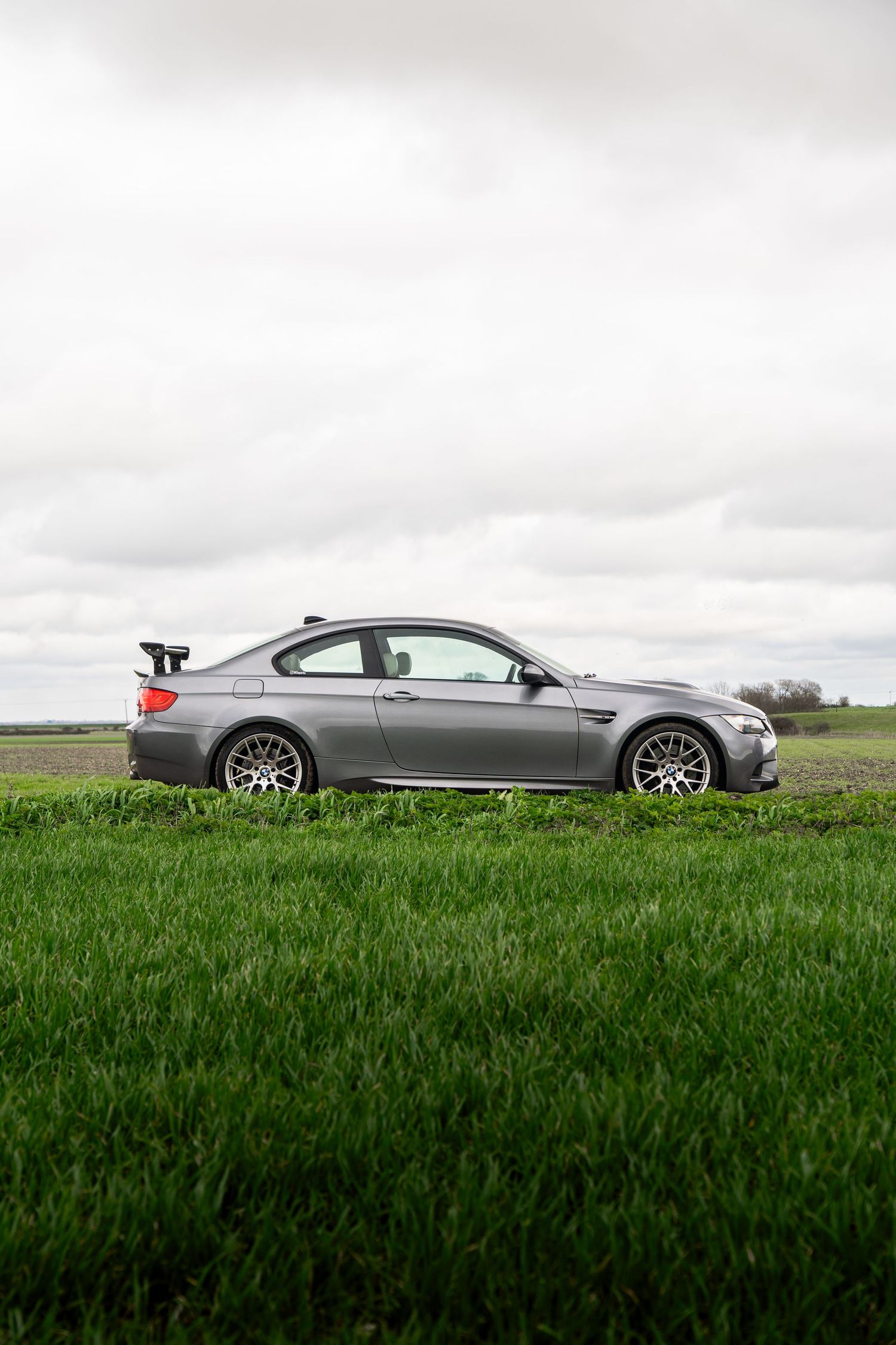 Side profile of grey BMW M3 coupe with racing spoiler on lush green grass field.