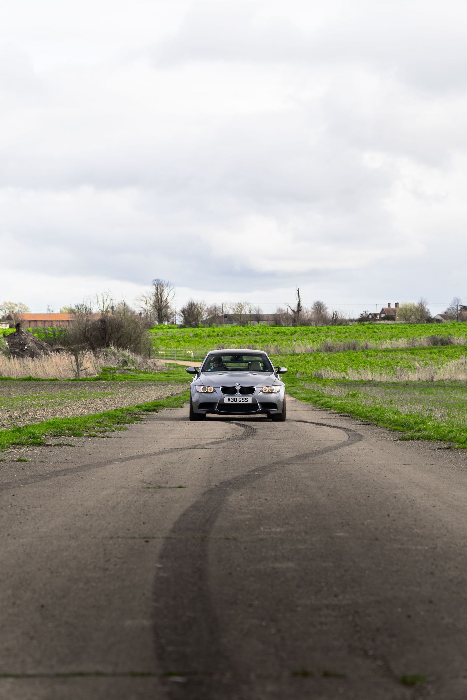 Silver BMW M3 driving straight on an empty rural road with tire marks visible.