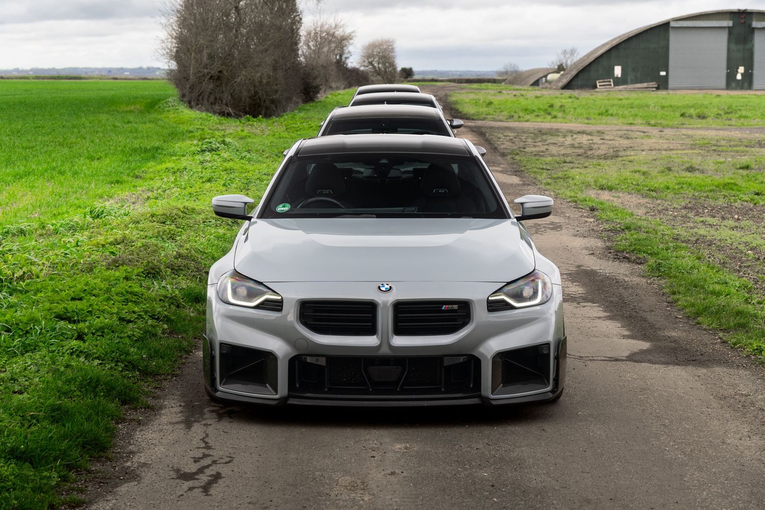Front-facing white BMW M2 Competition with carbon splitter on a rural dirt road.