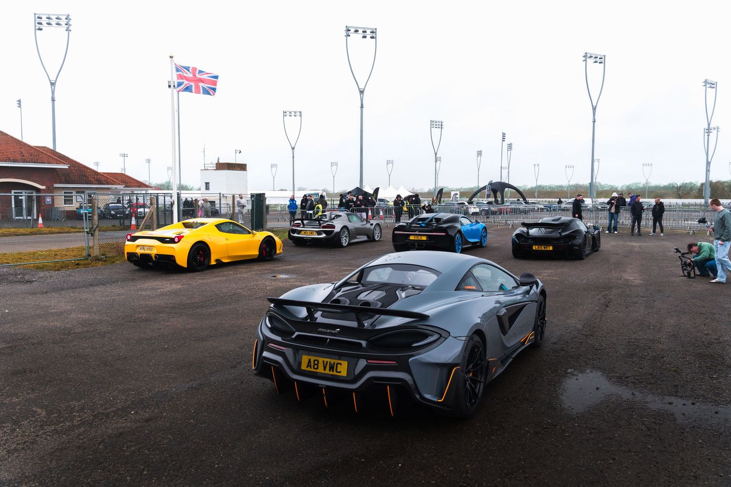 McLaren supercar leads lineup of exotic cars including Ferrari and Bugatti at UK track day event with Union Jack flag.
