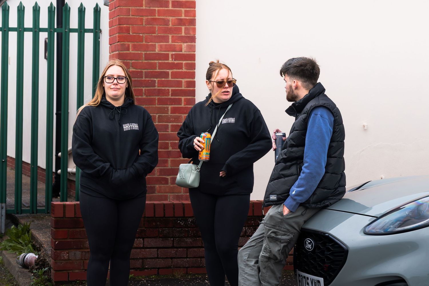 Two women and a man in casual black clothing chat outside near a brick wall and parked car.