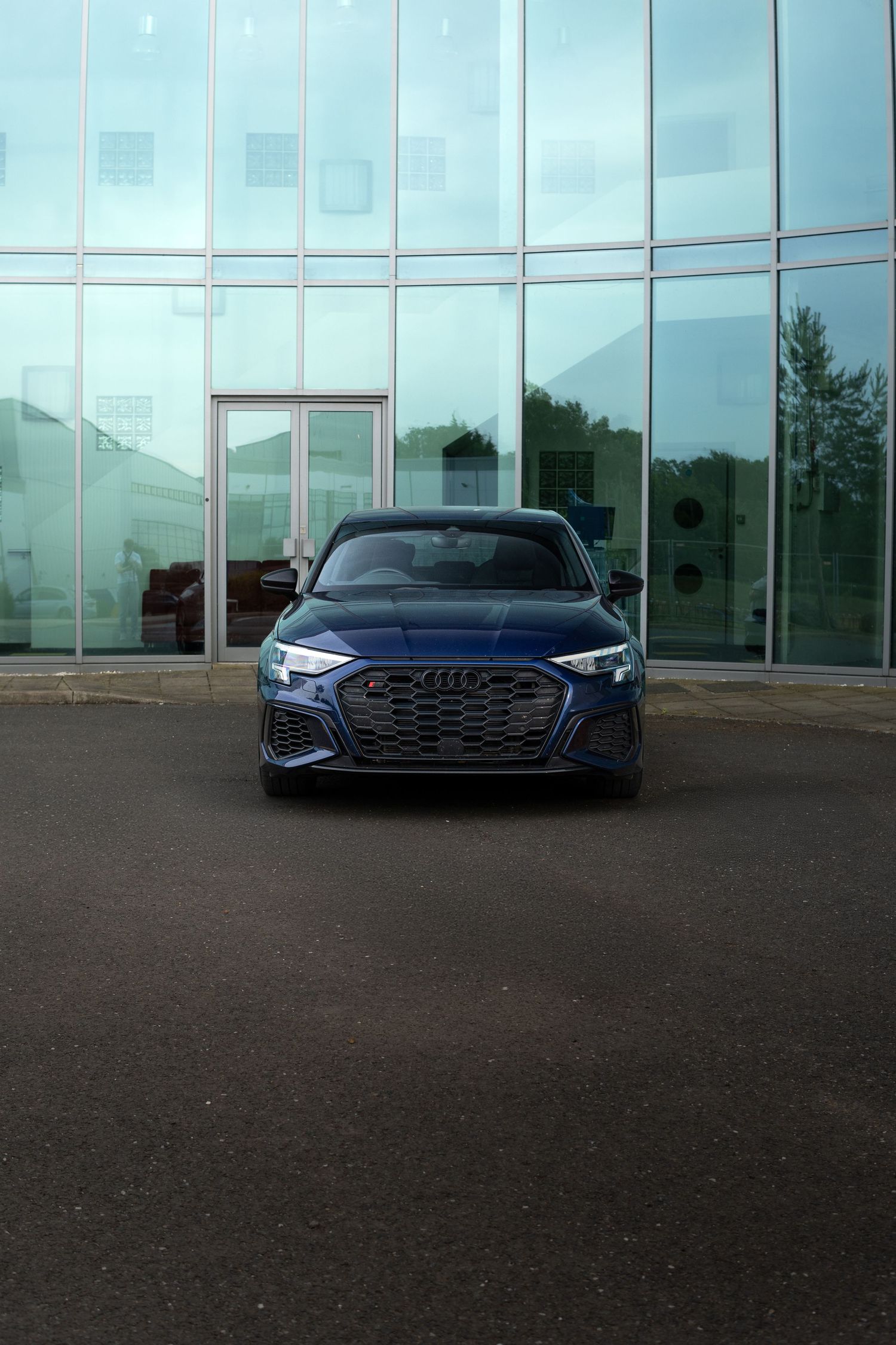 Head-on shot of a navy blue Audi S3 with aggressive grille in front of a glass facade.