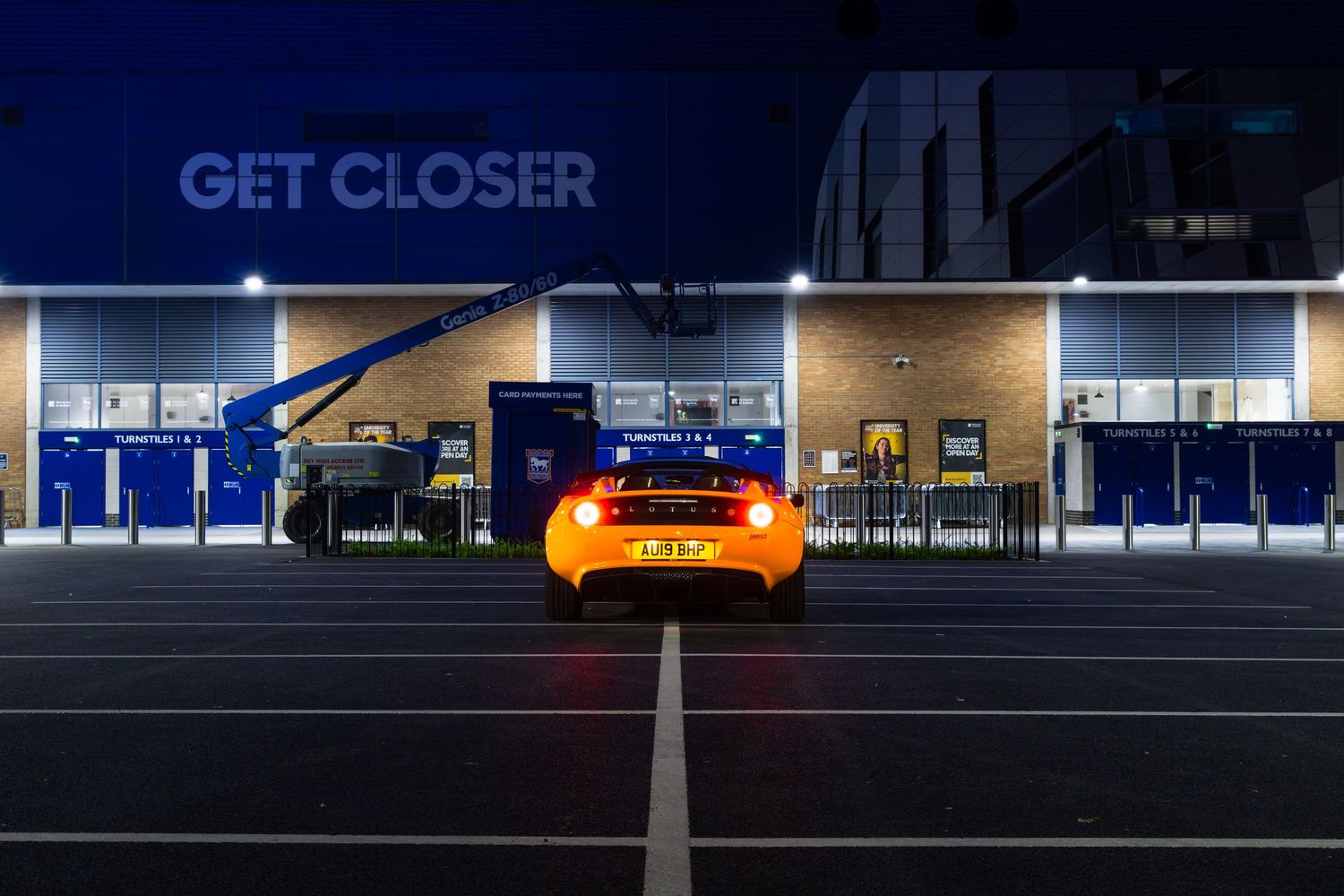 Rear view of orange Lotus Elise sports car at night outside illuminated GET CLOSER stadium entrance.