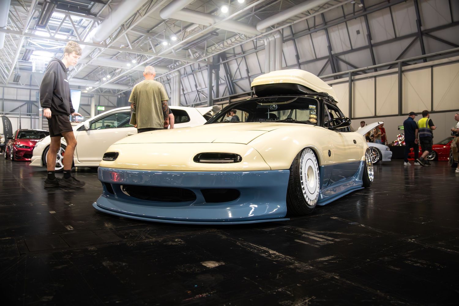 Heavily modified cream and blue Mazda MX-5 Miata with wide body kit on display at indoor car show.
