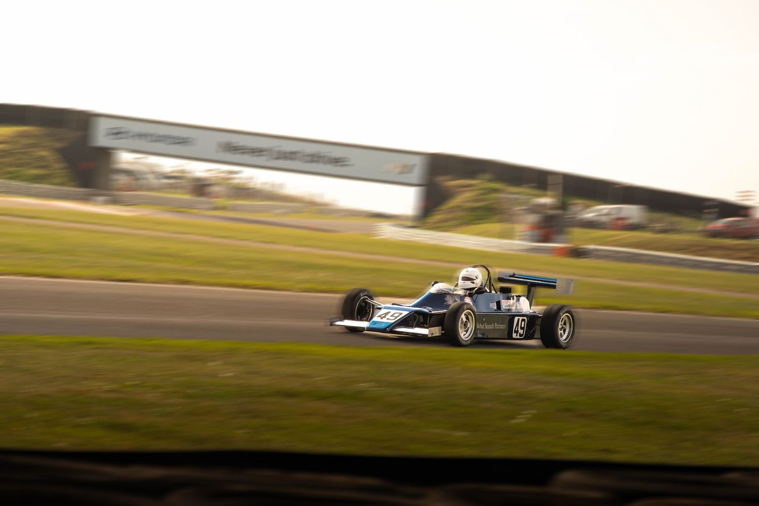 Classic blue and white Formula racing car speeds along a green grass-lined race circuit.