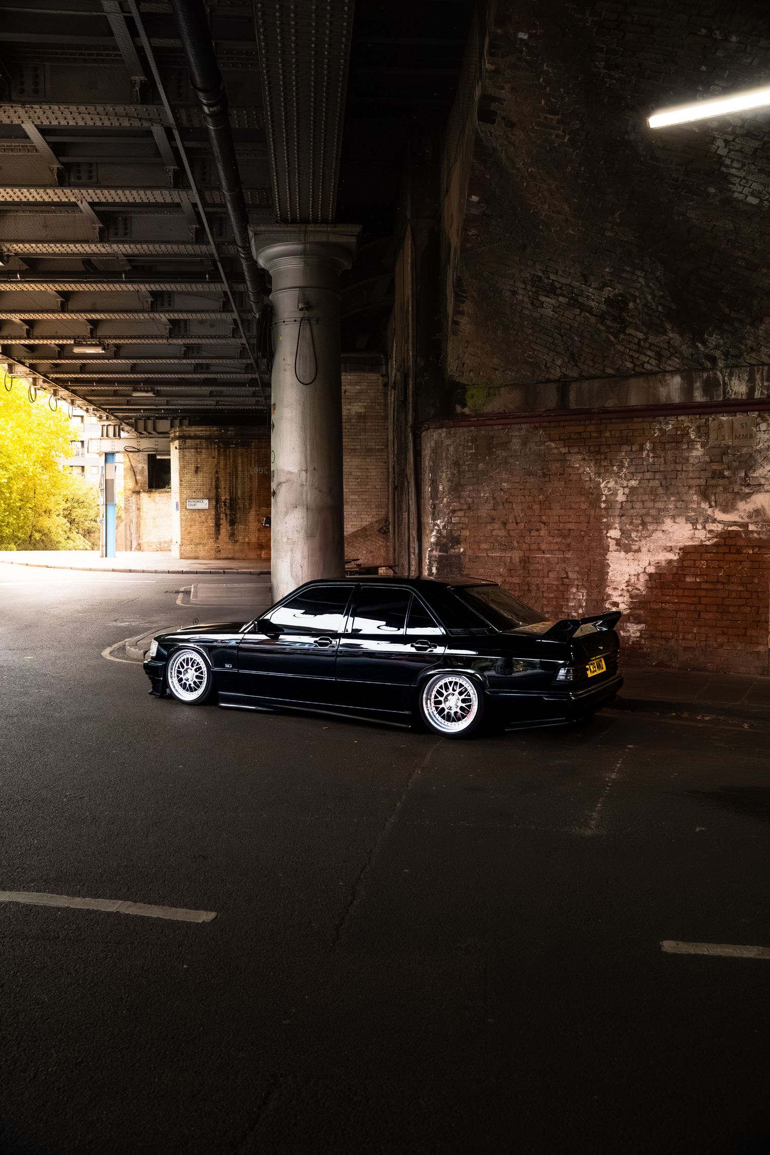 A slammed black BMW E38 on white BBS wheels parked under a dimly lit brick railway arch.
