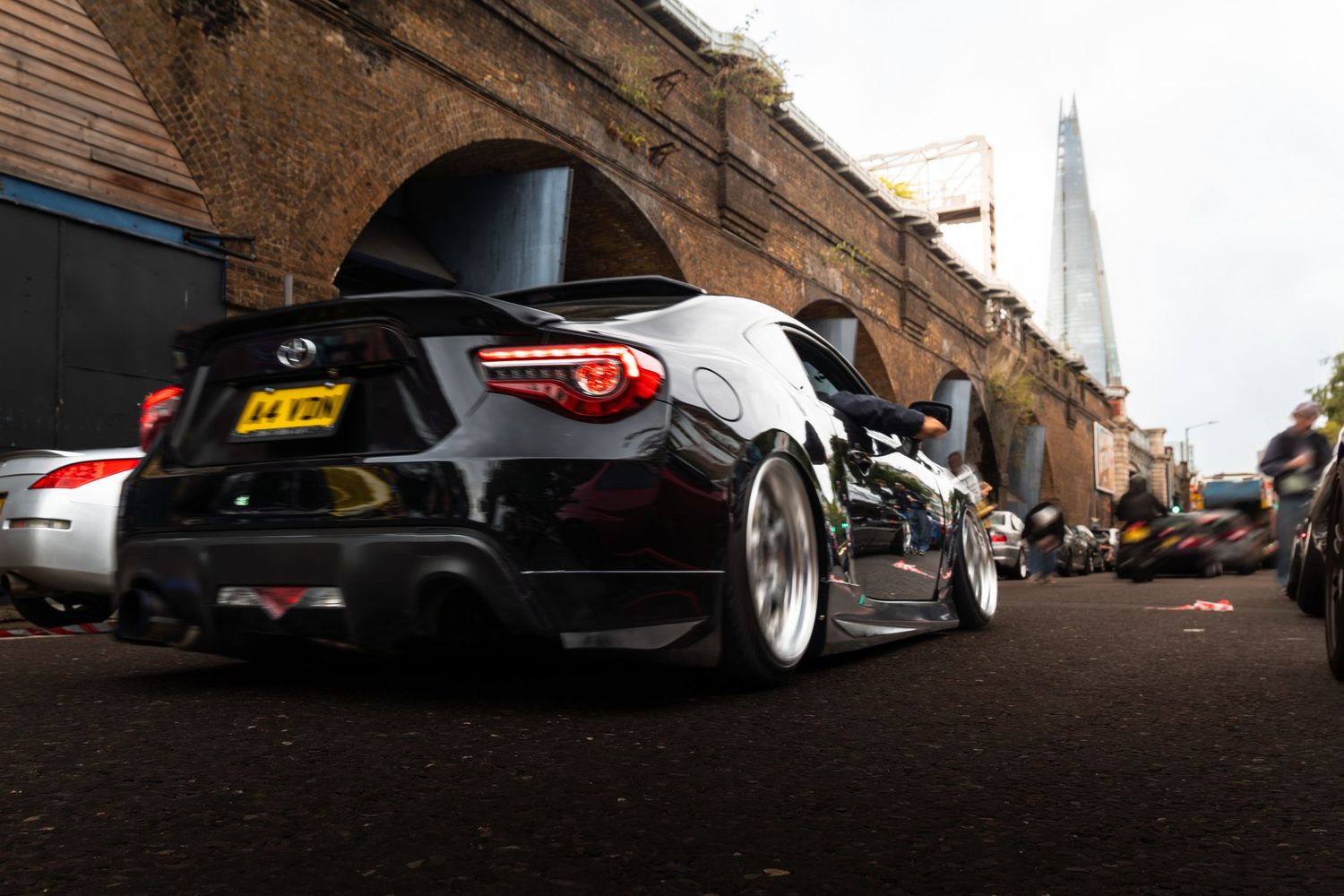 A black Toyota GT86 with wide body kit parked under a brick arch with The Shard visible.
