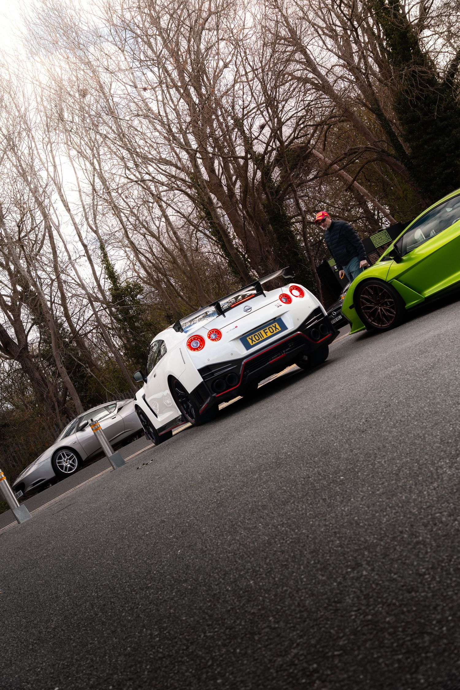 White Nissan GT-R Nismo and lime green McLaren parked on a road surrounded by bare winter trees at a car meet.