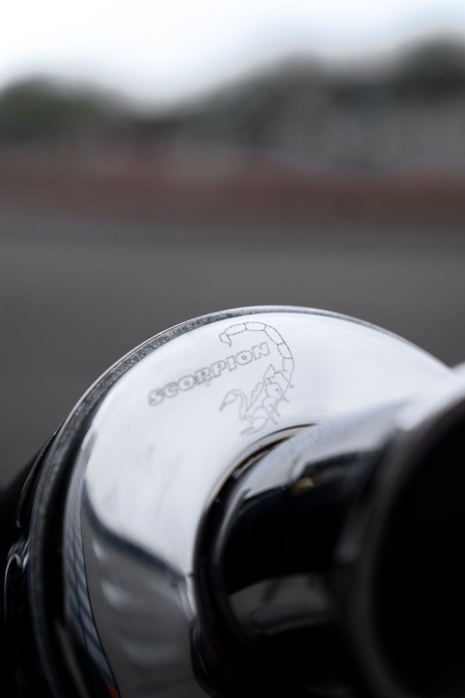 Chrome Scorpion exhaust end cap with engraved logo on a sport motorcycle at a racetrack.