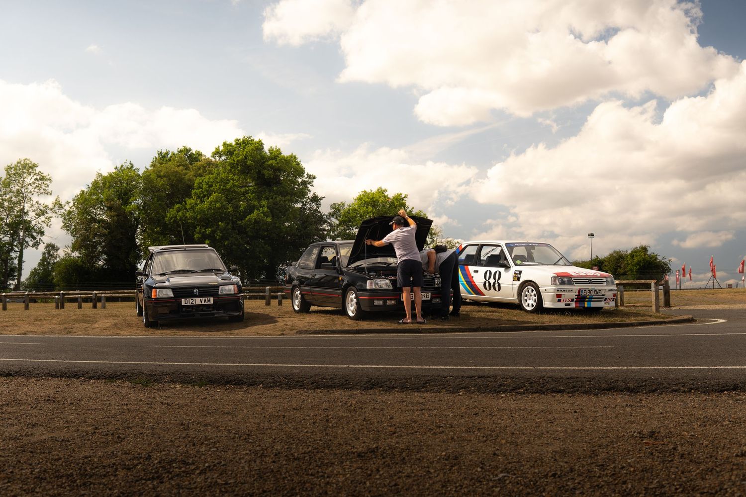 Three classic cars parked roadside with enthusiasts inspecting engines at an outdoor motorsport event.