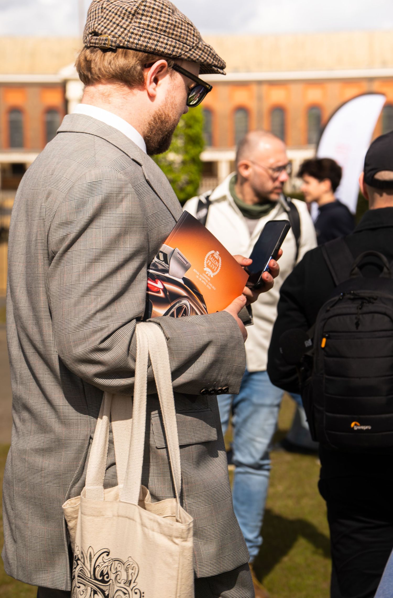 Stylishly dressed man in tweed jacket and flat cap reading an orange car show program.