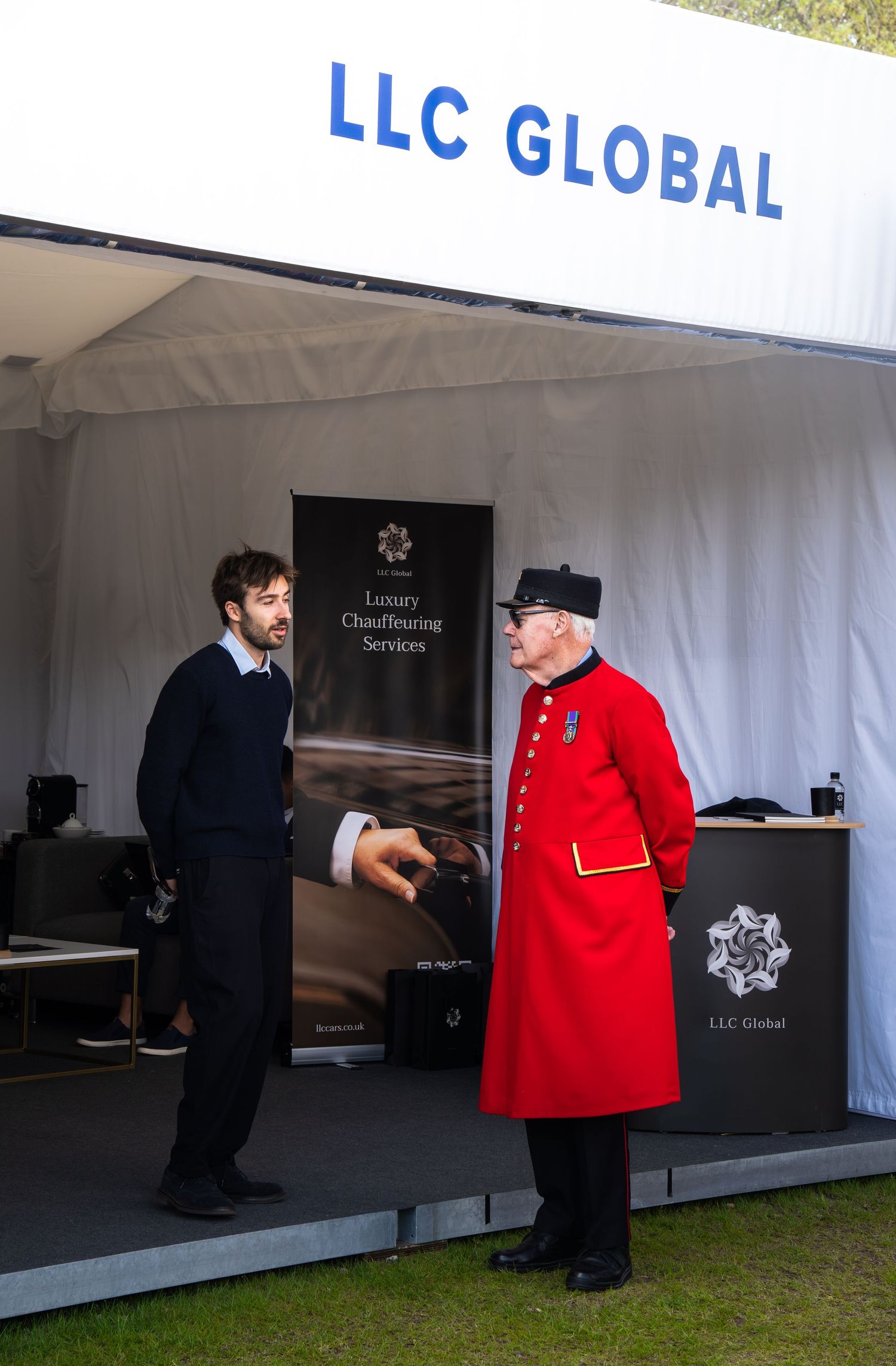 A Chelsea Pensioner in red uniform chats with a man at the LLC Global Luxury Chauffeuring Services booth.
