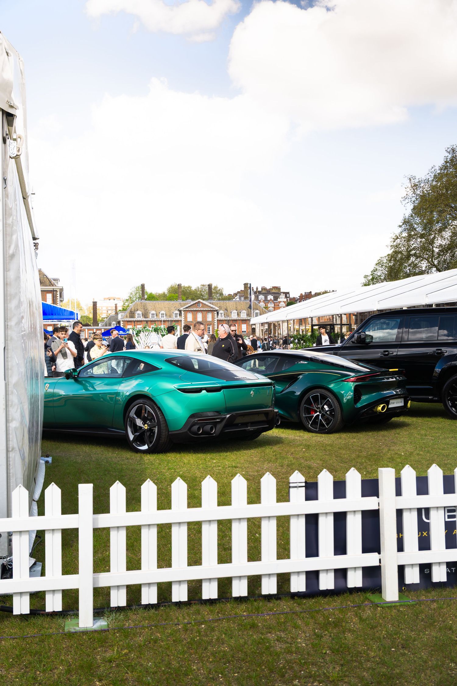 Two green Ferrari Roma and Lotus Emira sports cars displayed behind white picket fence at outdoor car show.